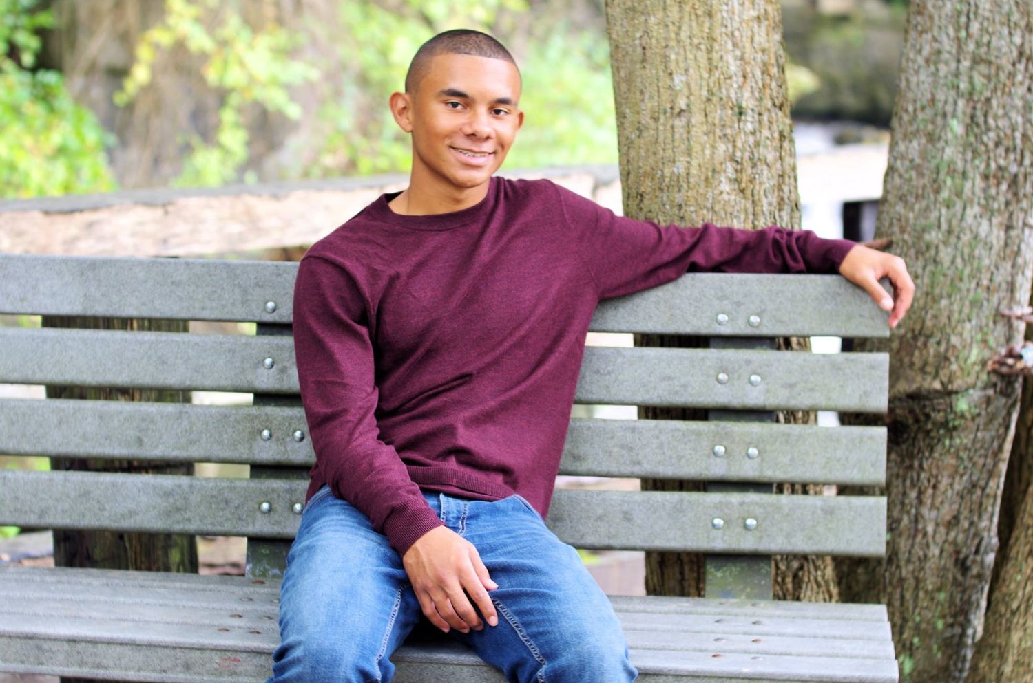 Boy in maroon sweatshirt sitting casually on wooden park bench.