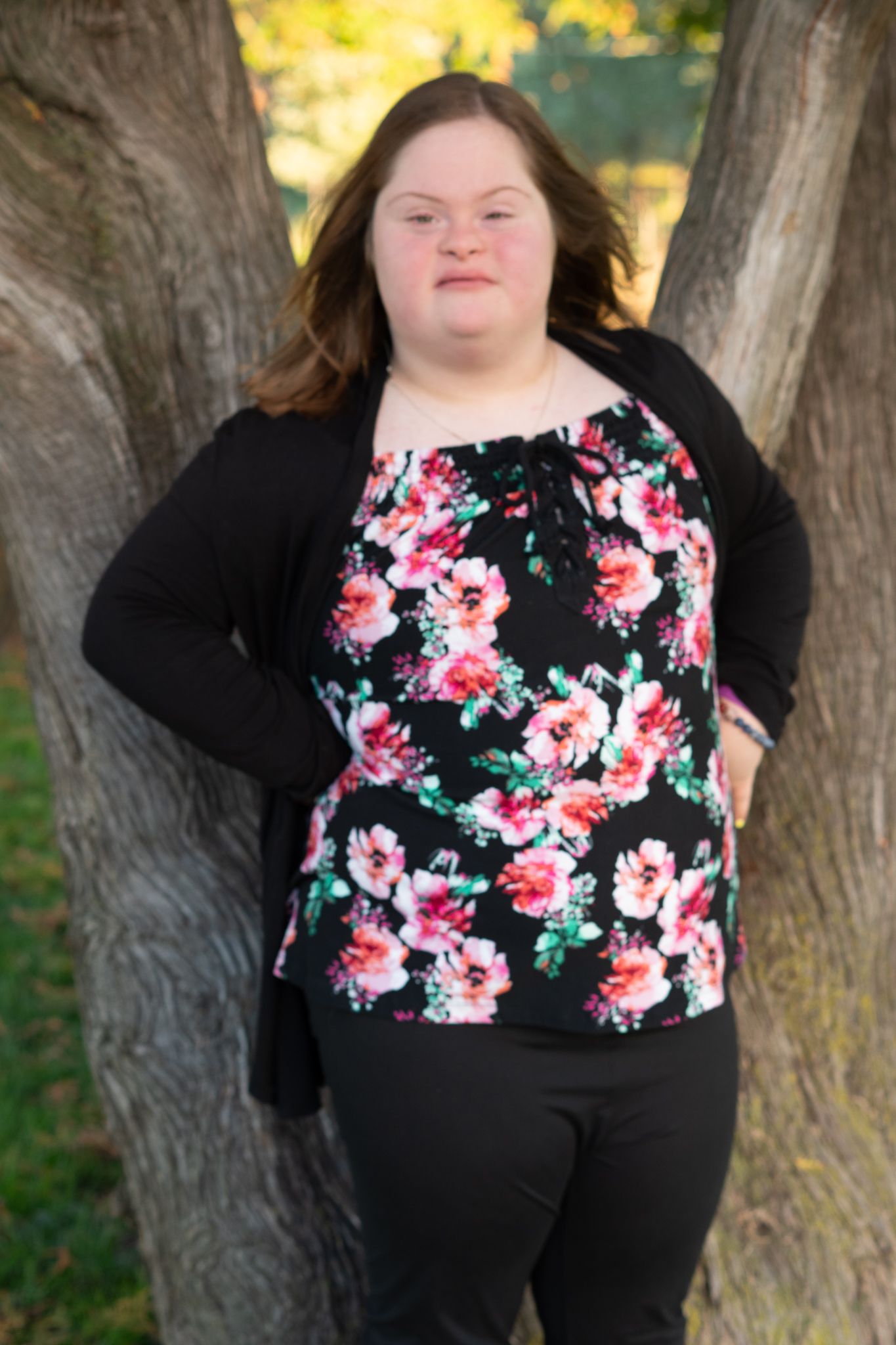 Young woman with Down syndrome in floral black top leaning against a tree outdoors.