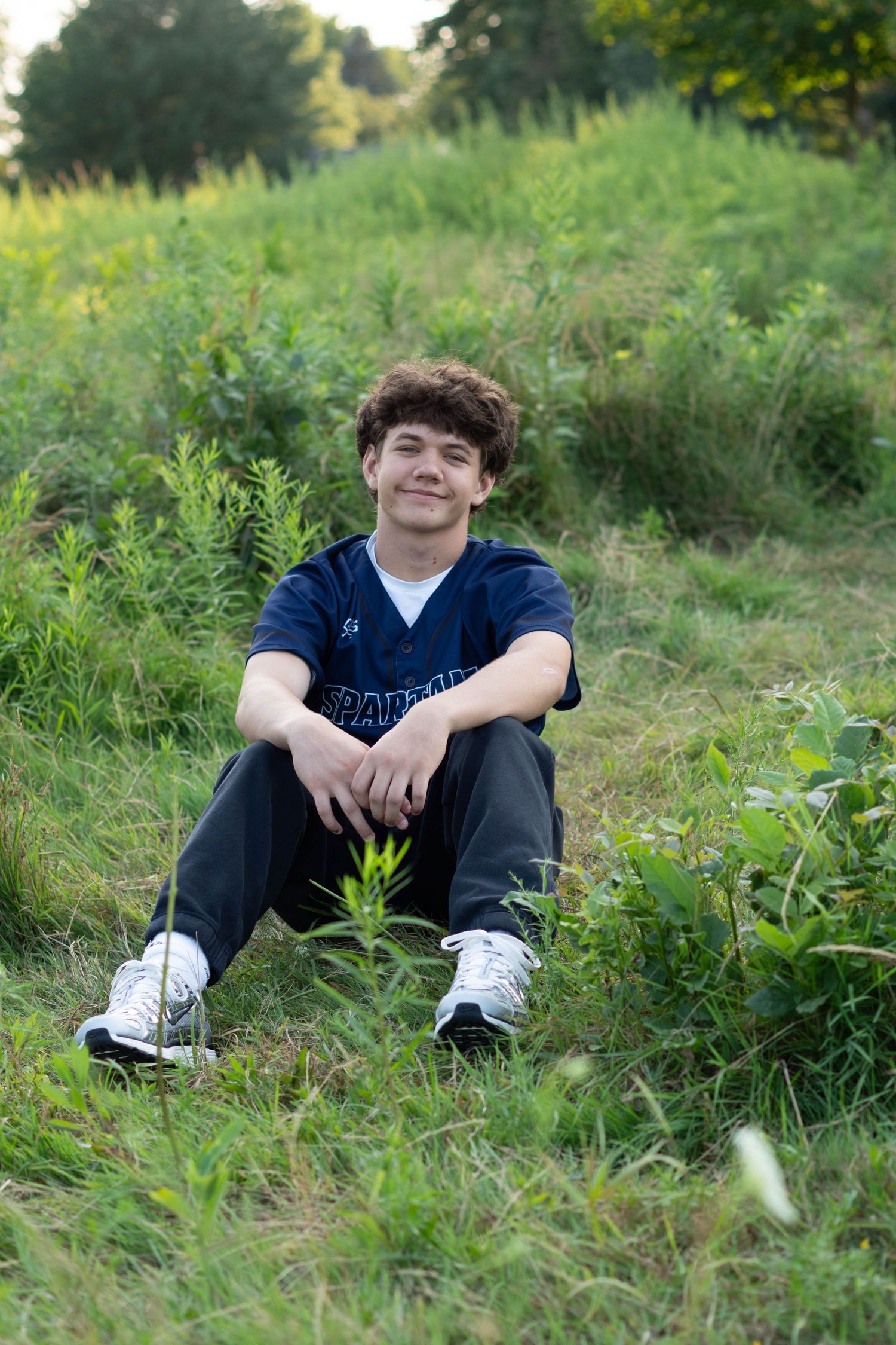 A smiling teenage boy sits cross-legged on green grass outdoors, wearing a navy blue shirt and dark pants.