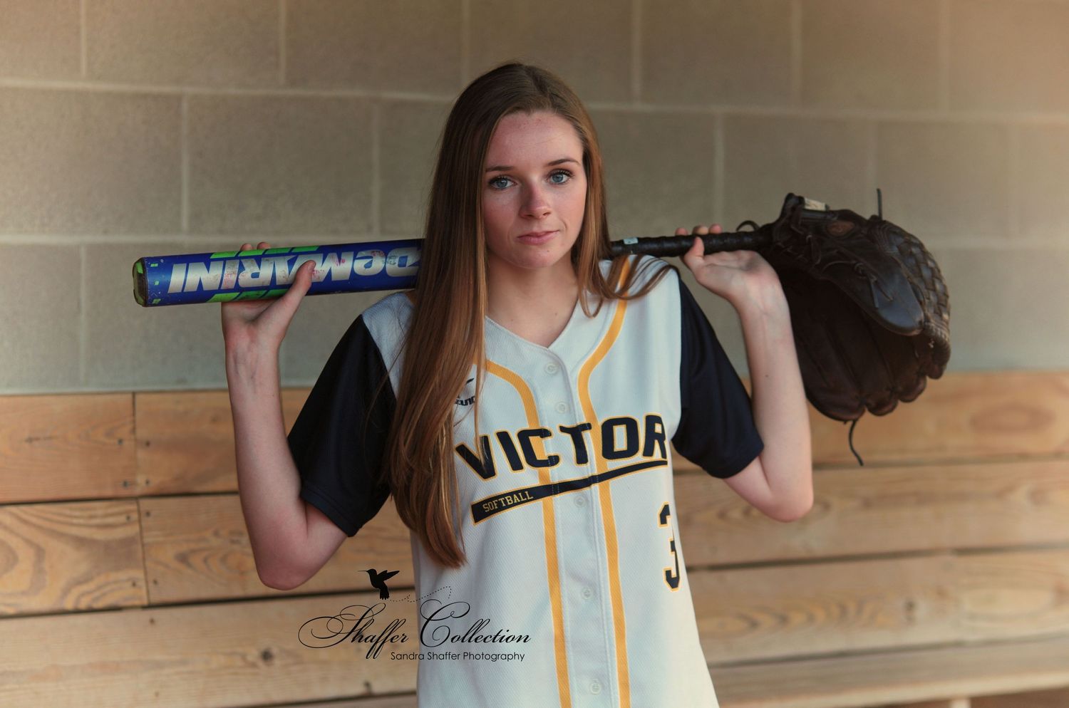 Softball player poses with bat and glove against beige wall wearing Victor team jersey.