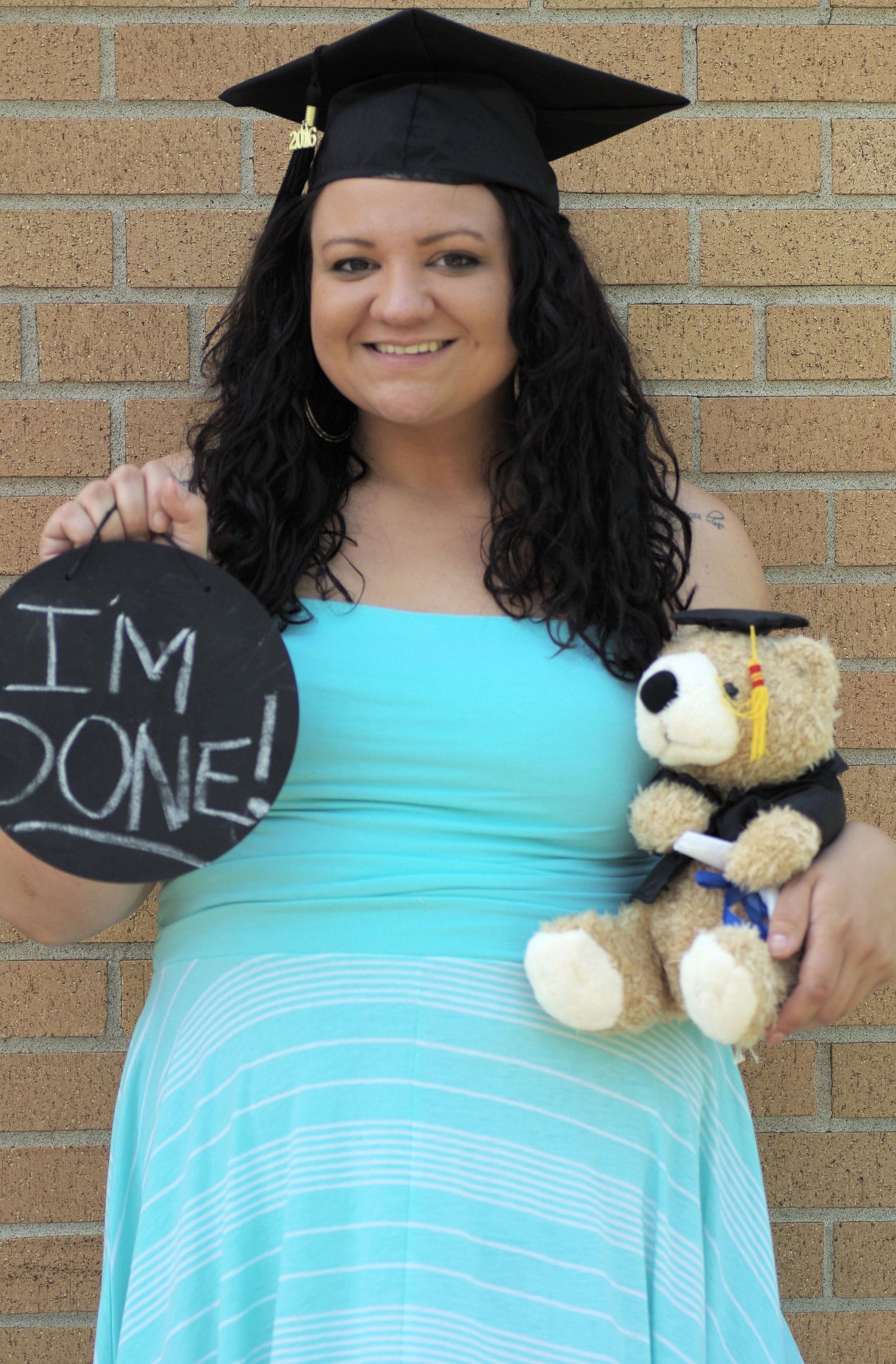 Graduate in cap and gown holds chalkboard sign and stuffed teddy bear against brick wall.