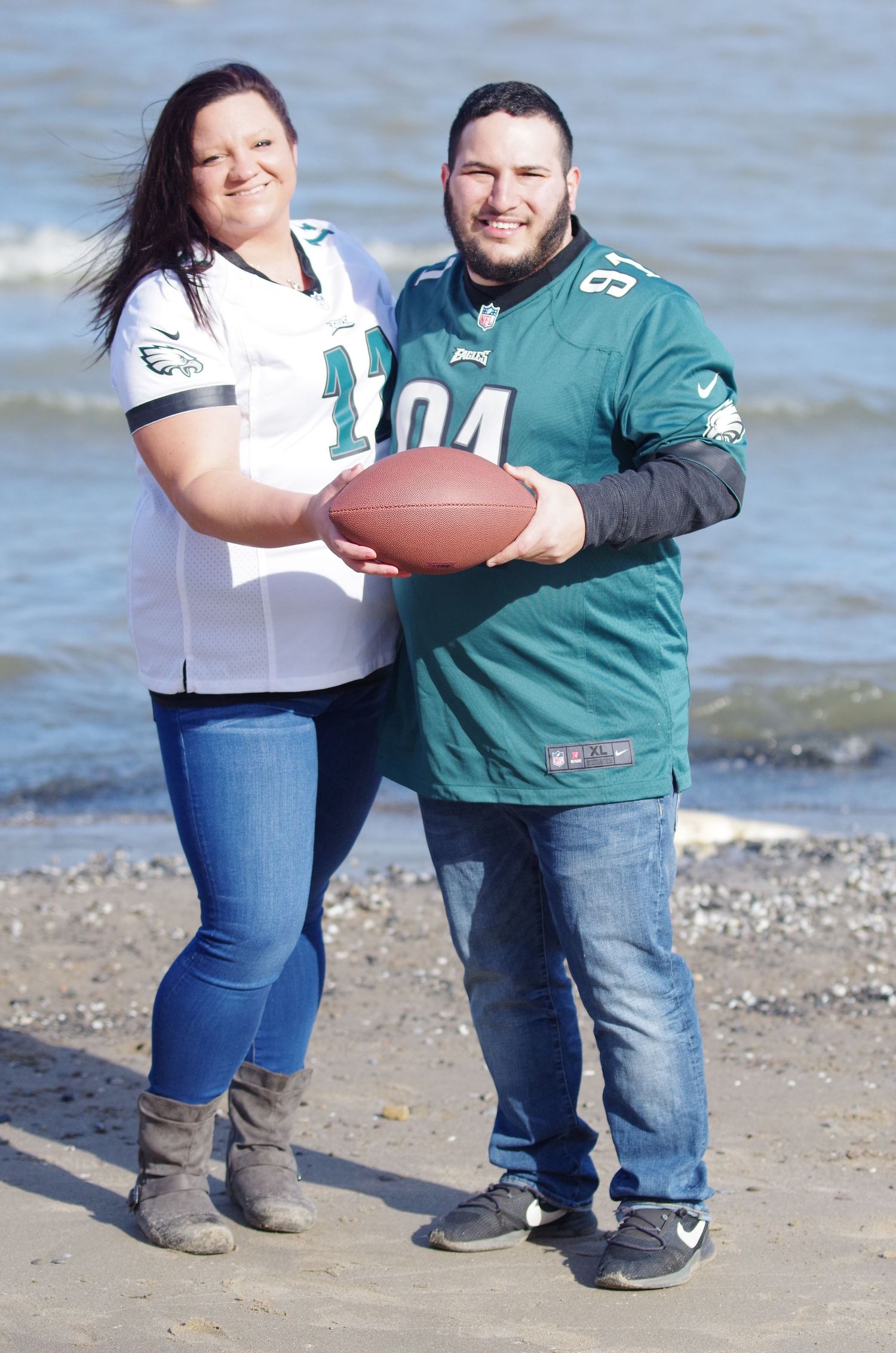 Couple in Eagles jerseys posing with football on beach.