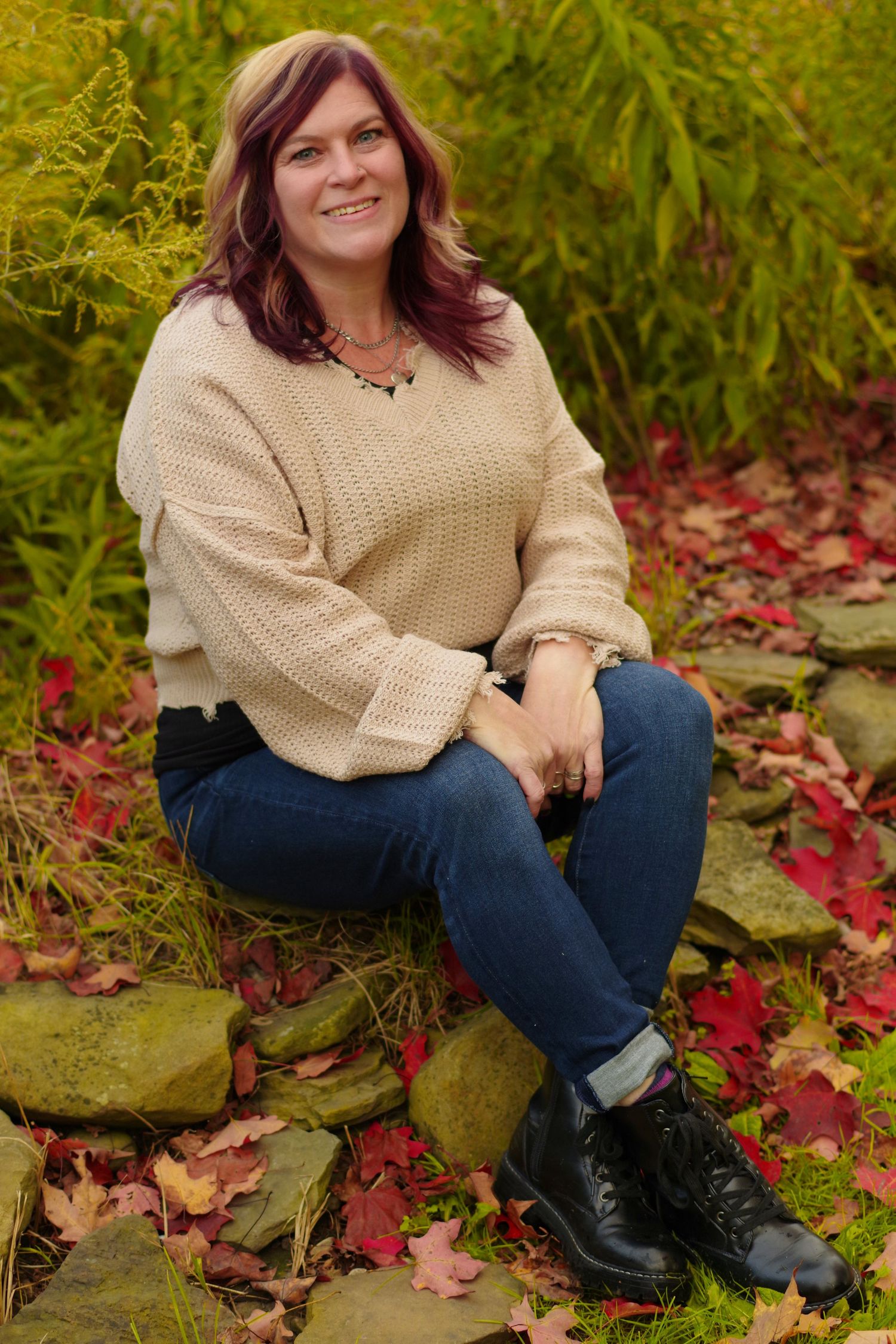 Person in beige sweater sits on stone wall surrounded by fall leaves and greenery.