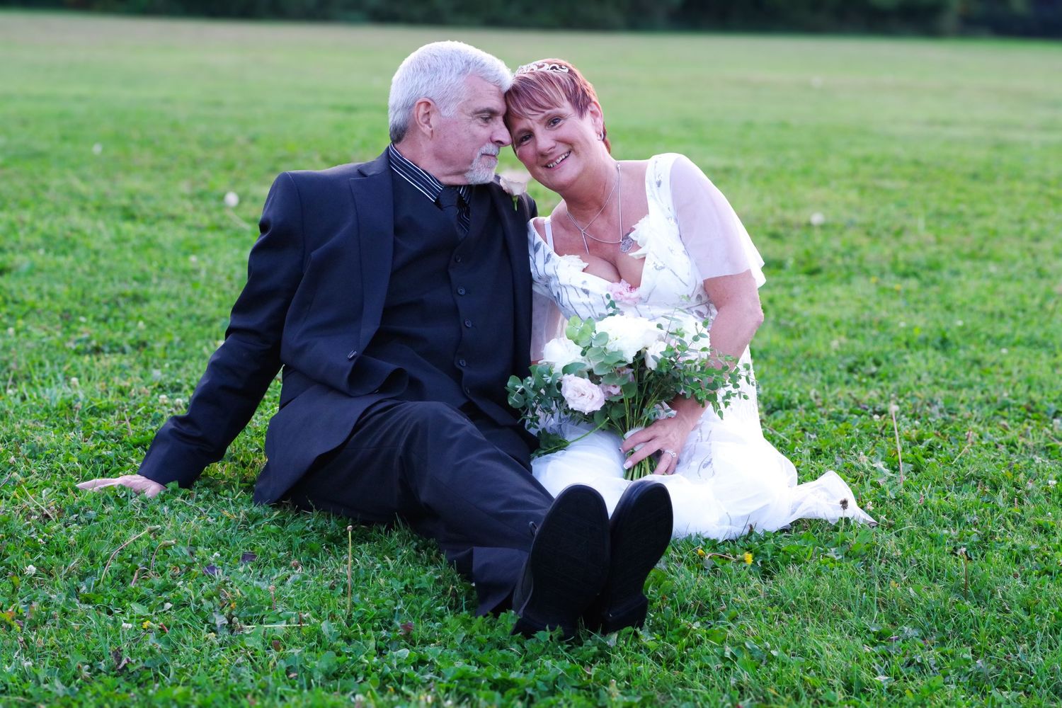 Wedding couple sitting on grass sharing tender moment with white floral bouquet.