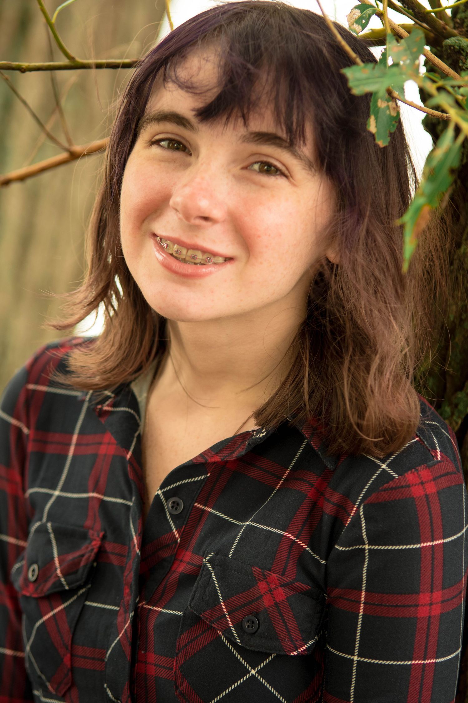 Person in plaid flannel shirt smiles while standing in front of green foliage.