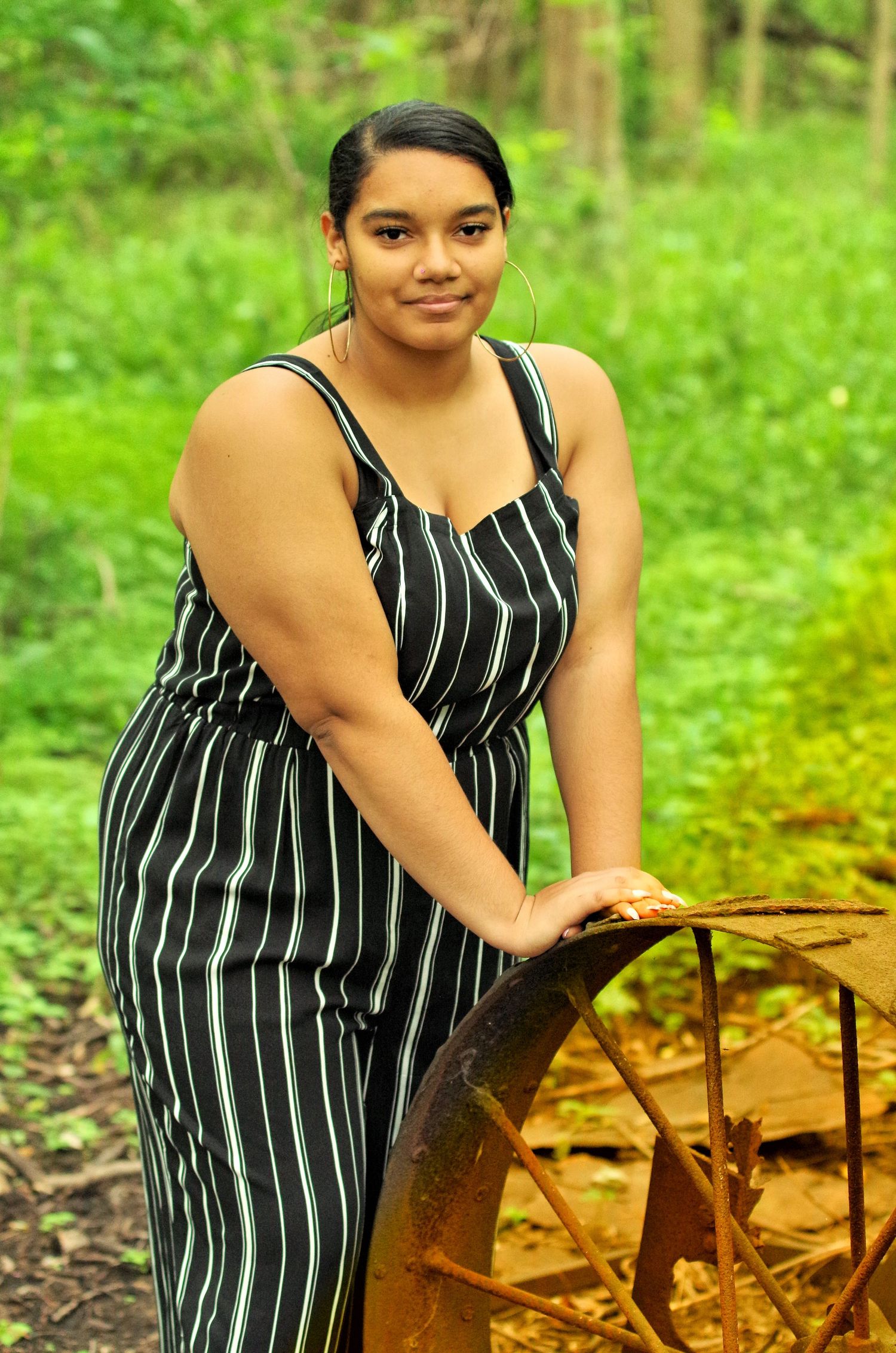 A summer portrait in a striped dress next to vintage farm equipment in a wooded setting.