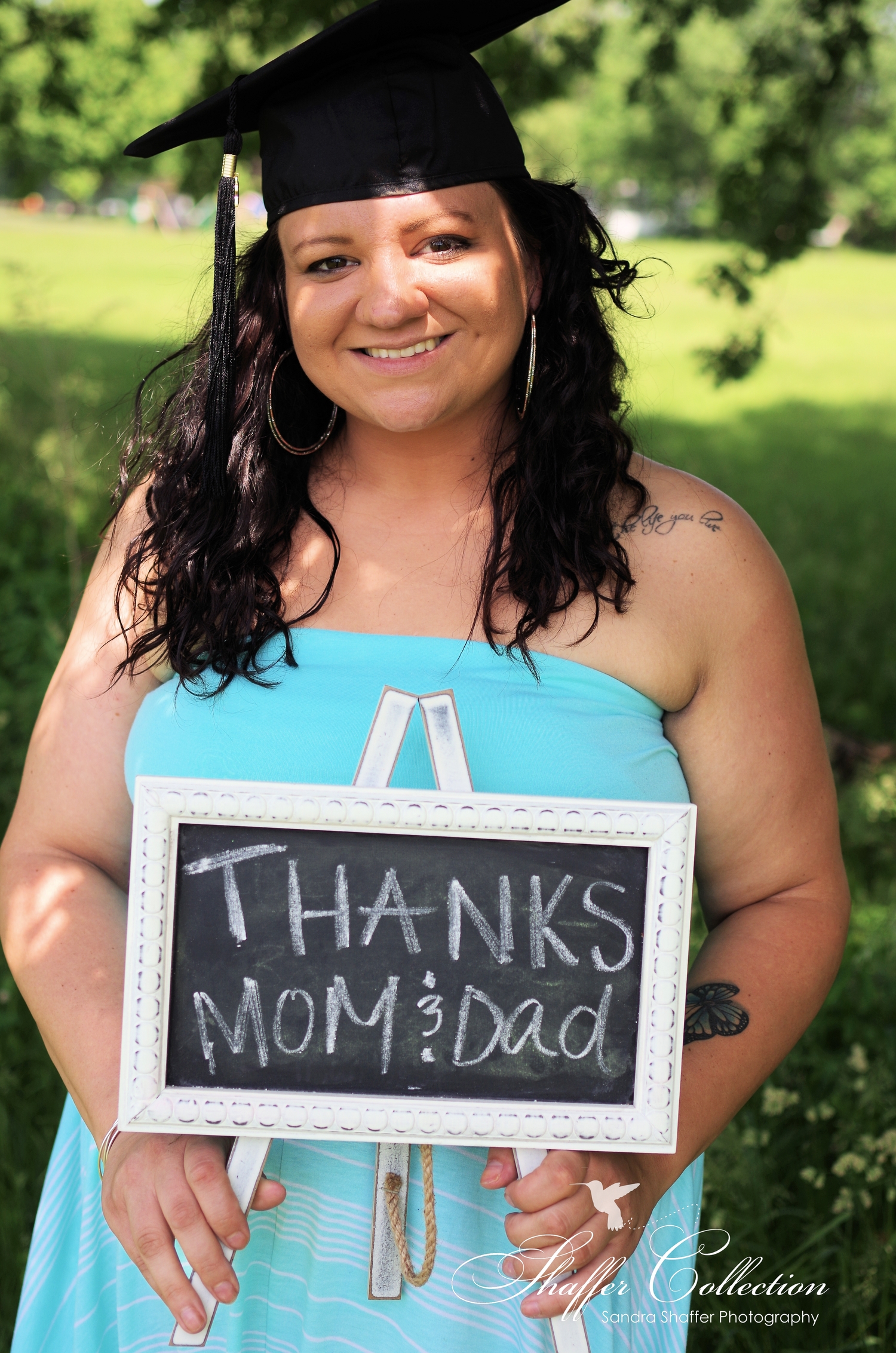 Graduate in blue dress holds chalkboard sign thanking parents during outdoor photoshoot.