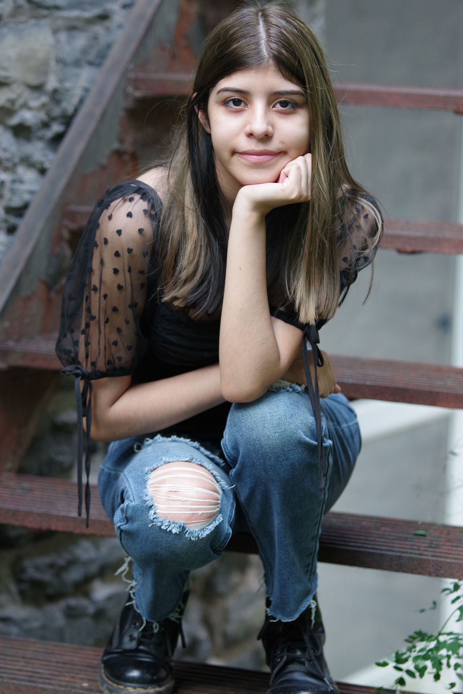 Casual portrait on steps wearing sheer black top and ripped jeans with hand on chin.