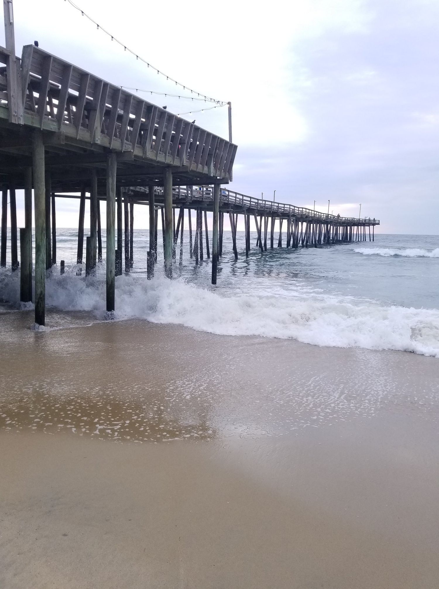 Waves crash beneath a wooden fishing pier extending into the ocean on a cloudy day.