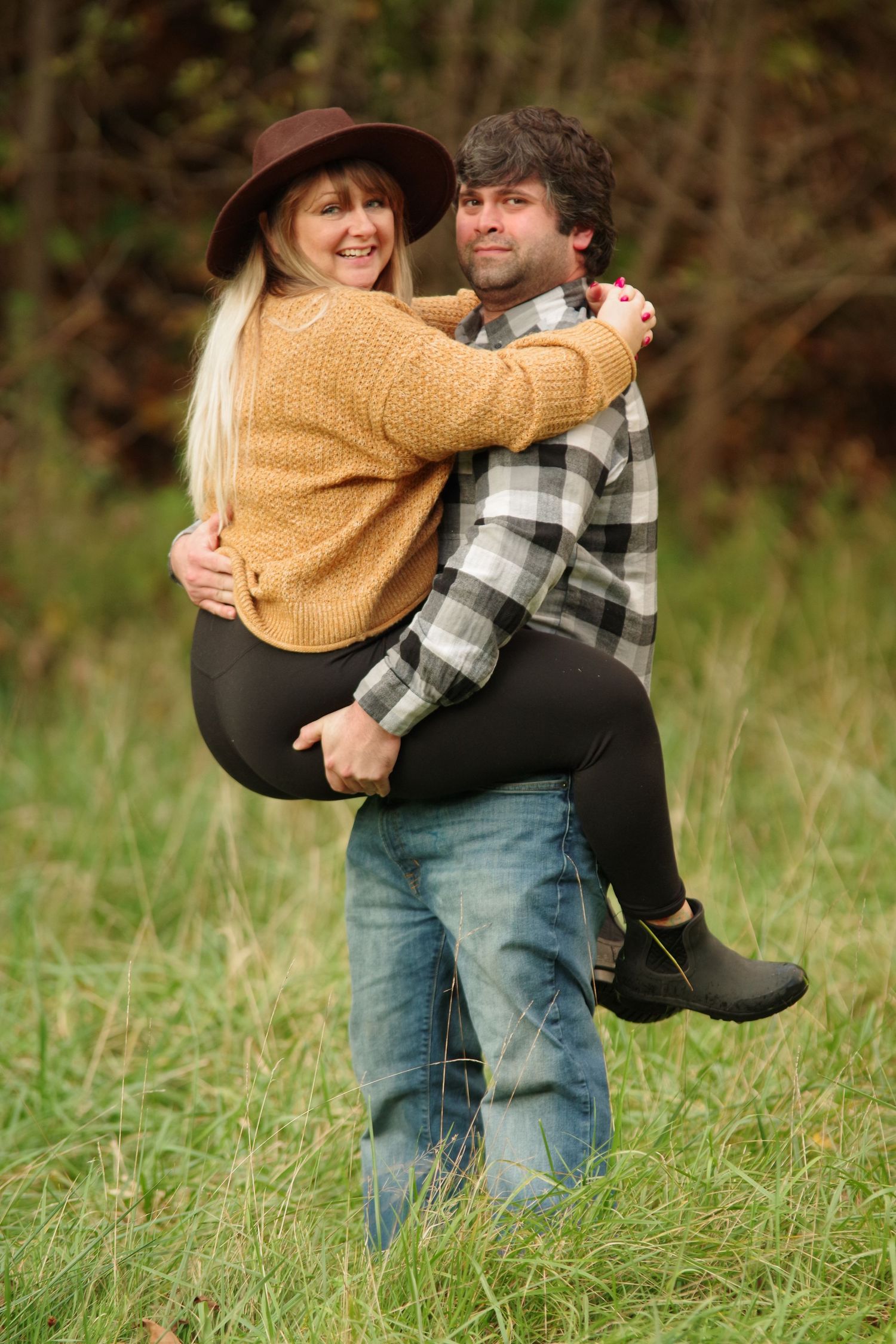 A man in a plaid shirt carries a smiling woman in a yellow sweater and brown hat outdoors in a grassy field.