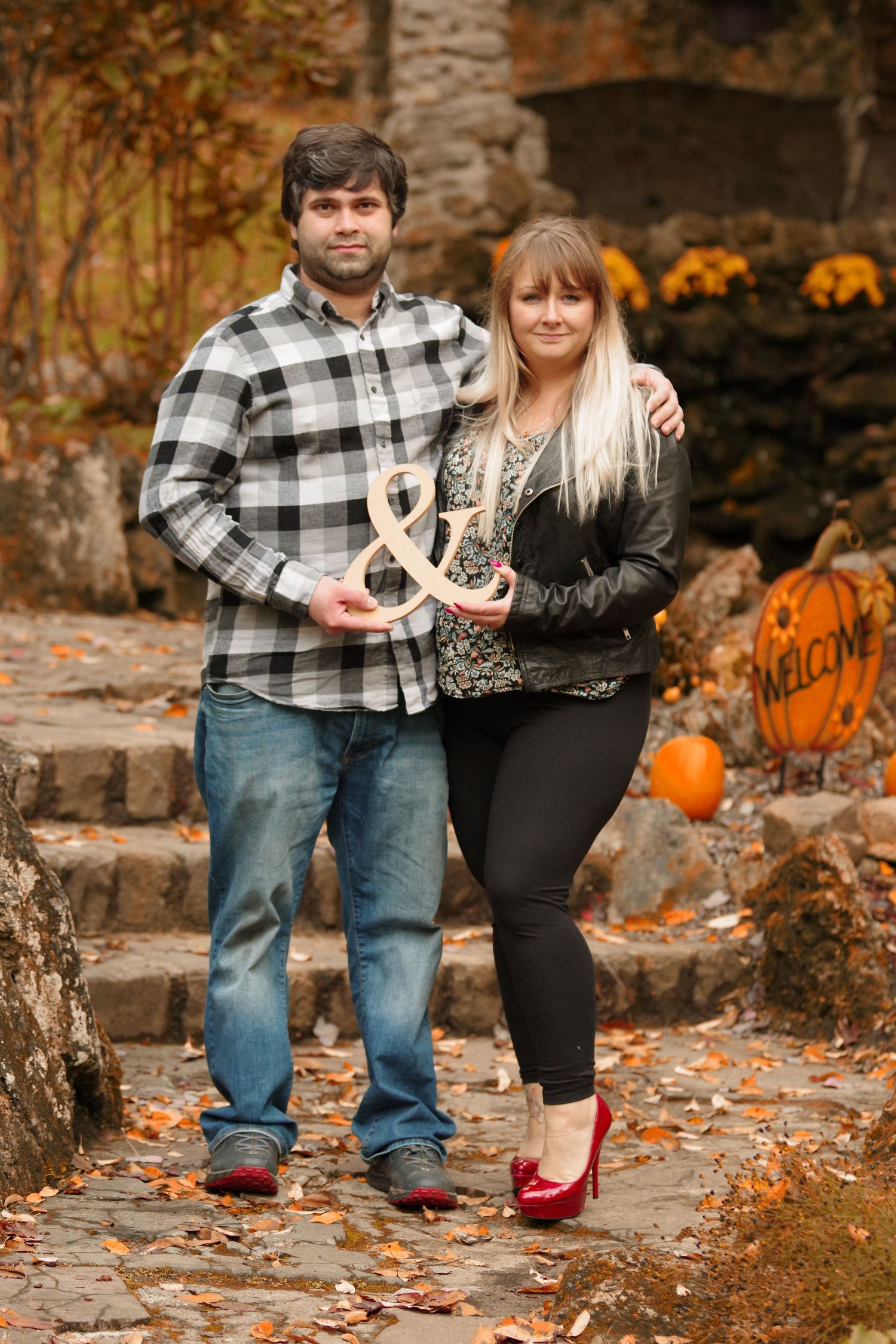 Couple posing together in autumn scene with pumpkins and fallen leaves on stone steps.