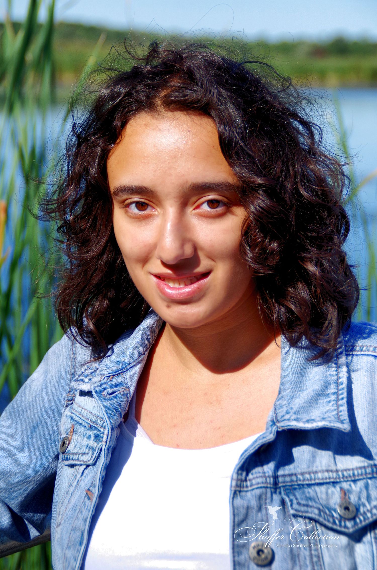 Natural sunlit portrait featuring casual denim jacket and flowing dark hair by a lakeside with reeds.
