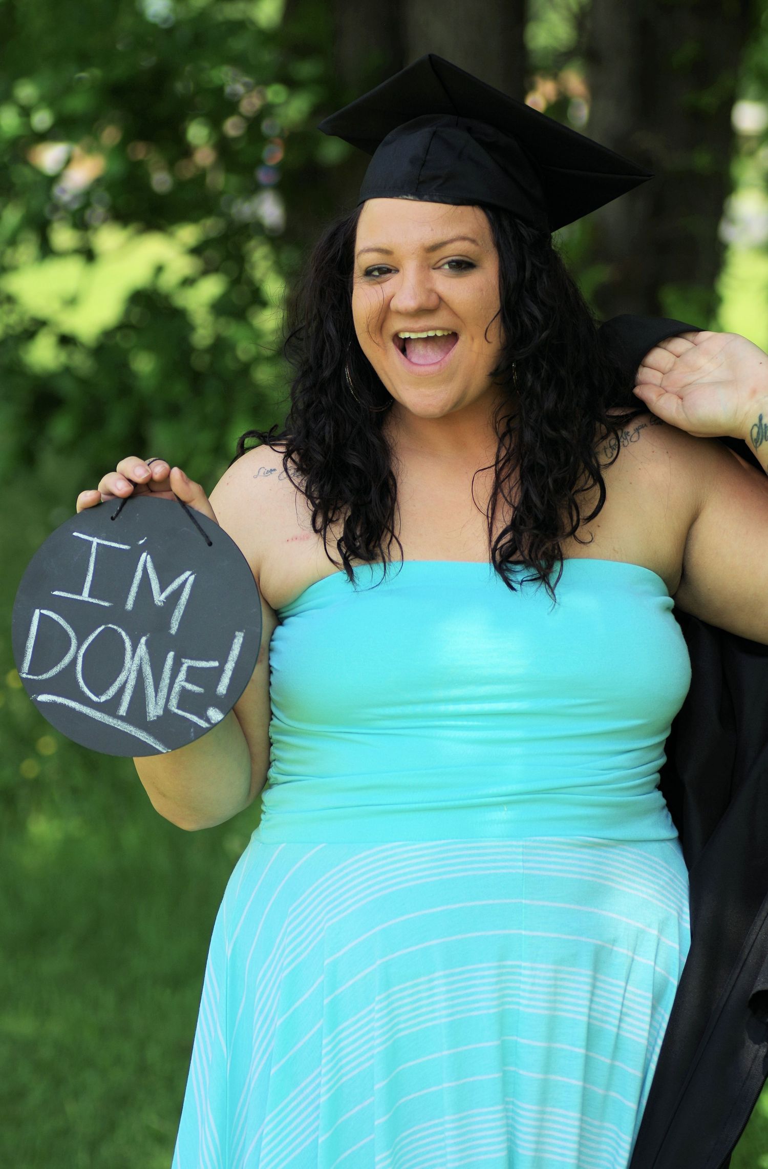 A smiling graduate in a teal dress and cap holds a chalkboard sign reading I'm Done outdoors.