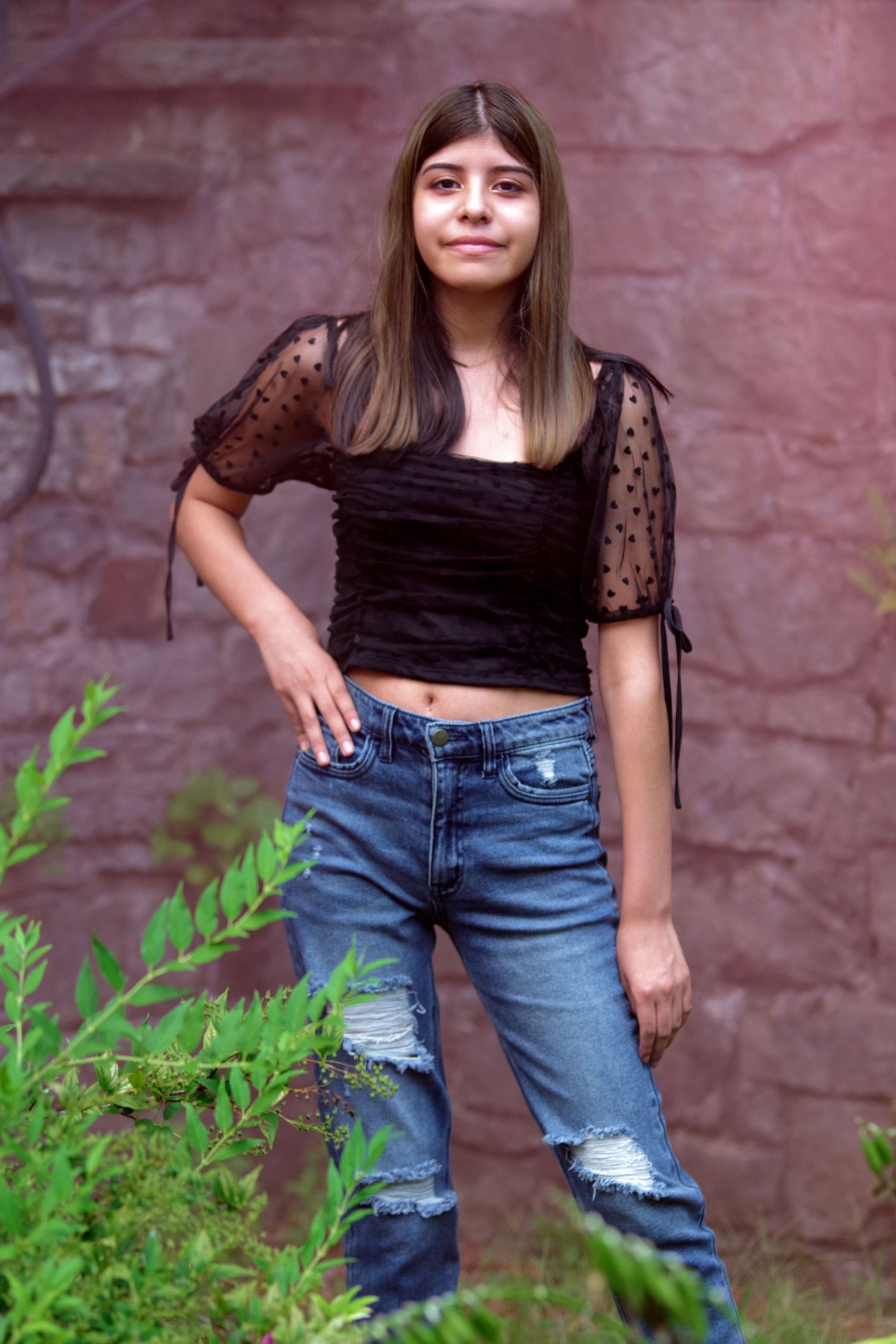 Fashion portraits showing distressed denim and black top with greenery and stone wall backdrop.