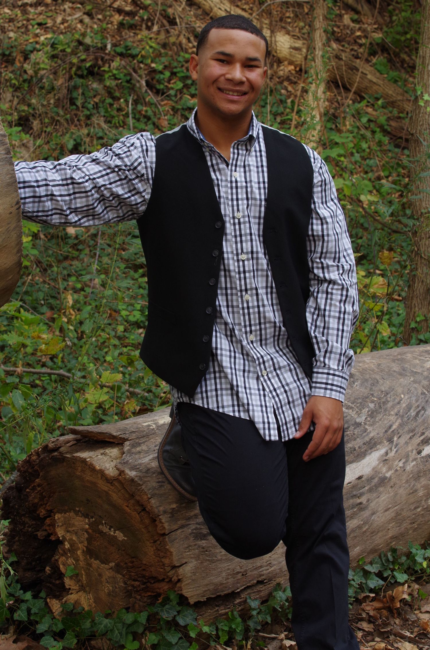 A person in a checkered shirt and black vest sits casually on a fallen log in a forest setting.
