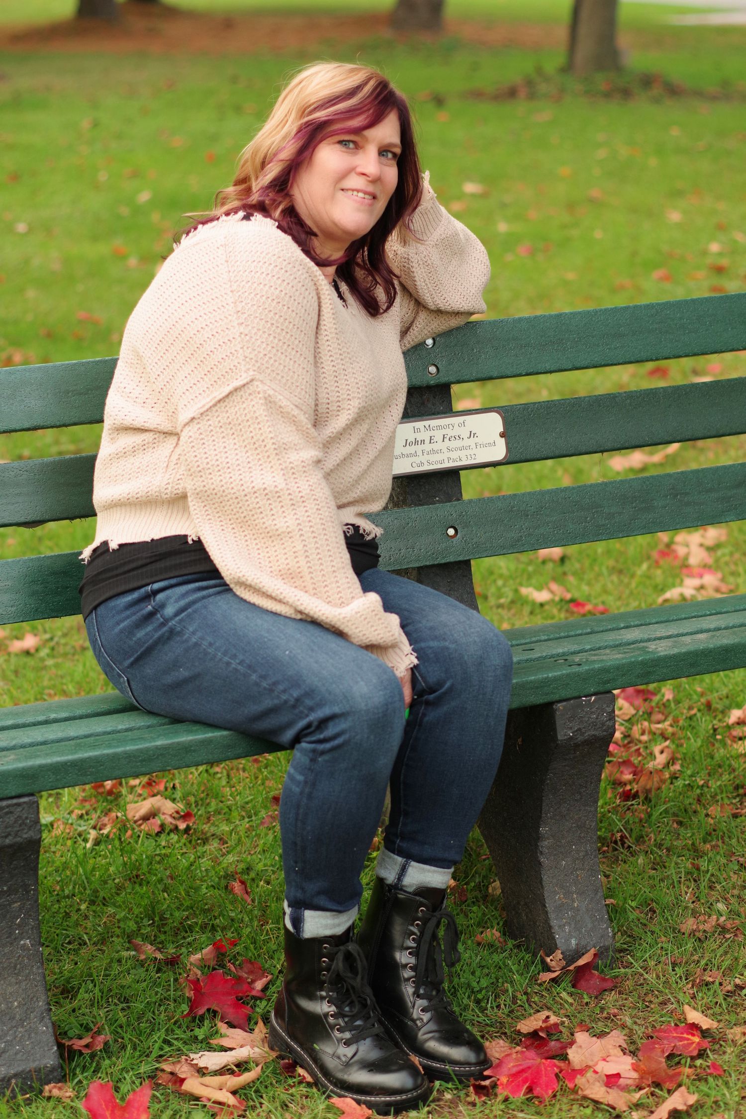 Person in cream sweater and jeans sits casually on a green park bench surrounded by autumn leaves.