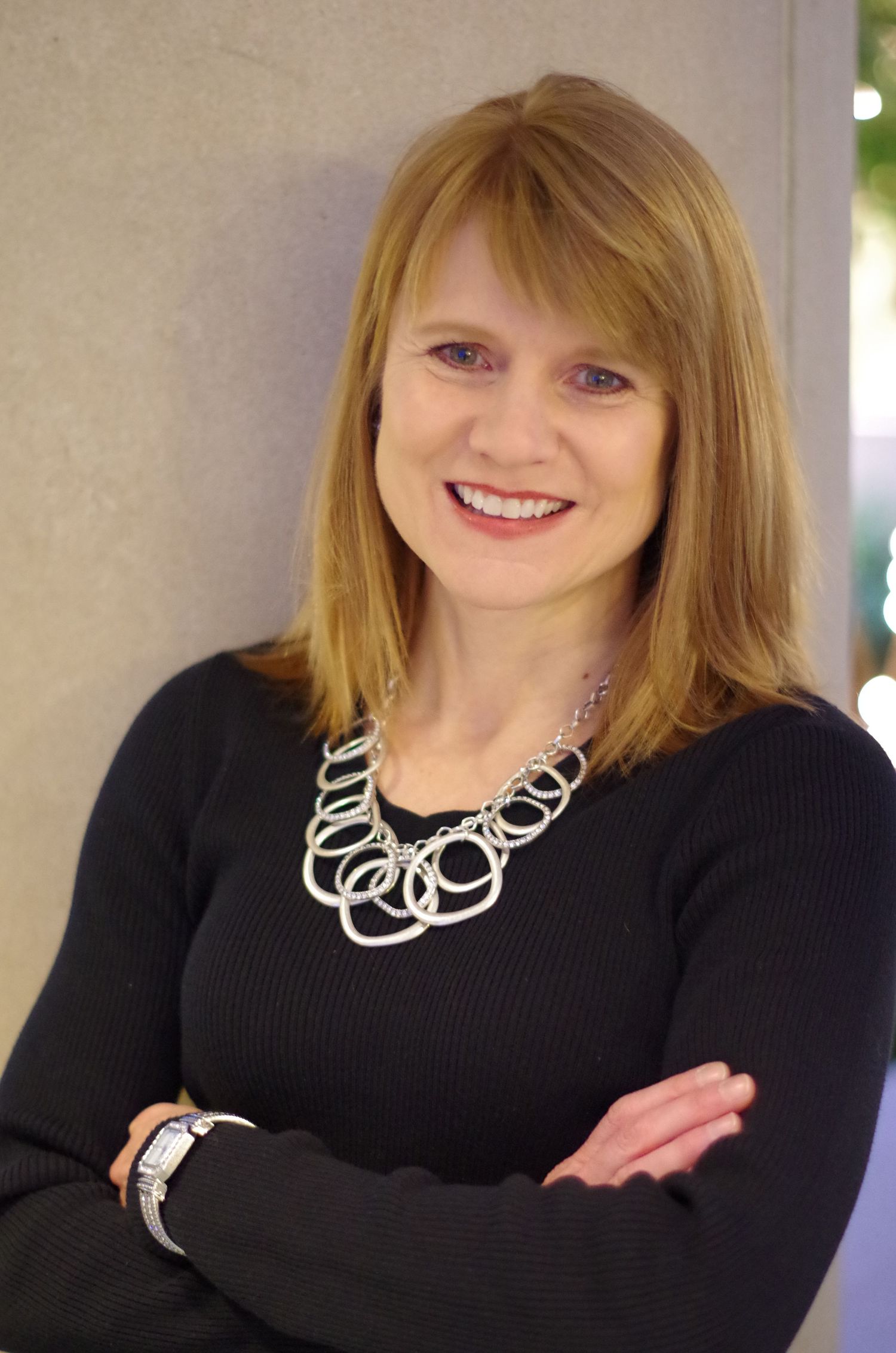 Professional headshot with black sweater and silver circular chain necklace against light background.