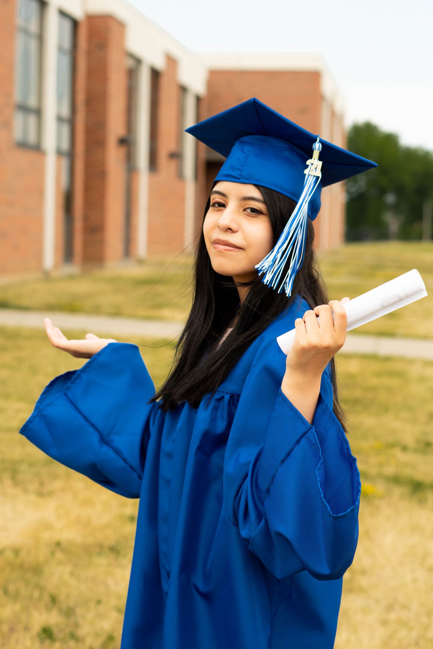 Graduate in blue cap and gown poses playfully with diploma outdoors.