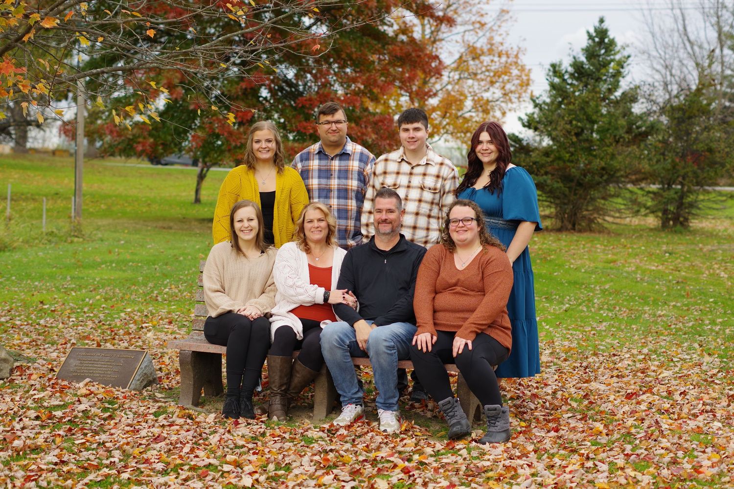 Family of eight posing outdoors in autumn, surrounded by colorful fallen leaves under a tree.