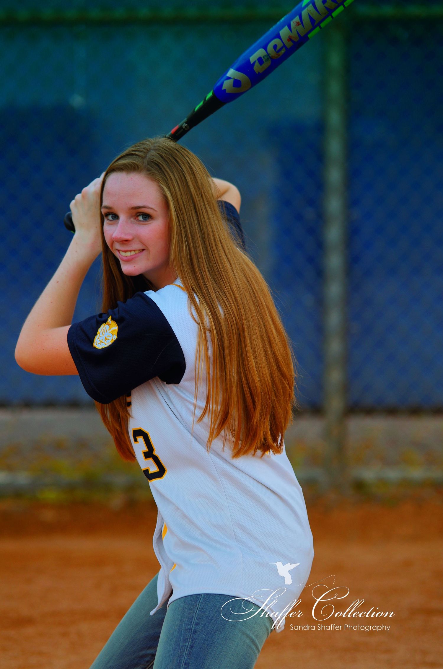 Softball player poses with blue and purple bat on dirt field during golden hour.