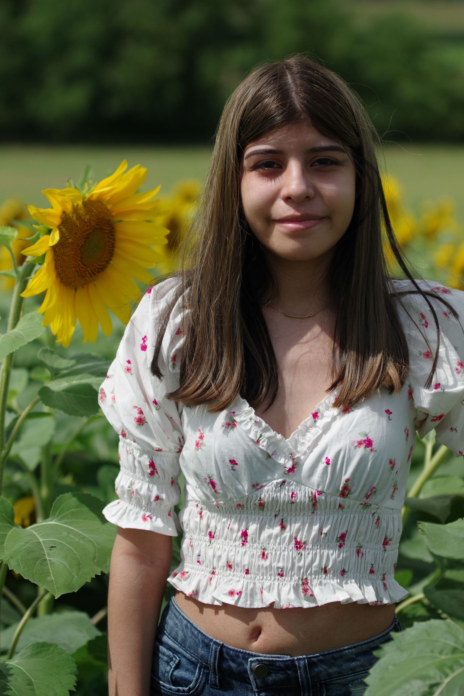 A person poses in a floral crop top in front of bright yellow sunflowers on a sunny summer day.