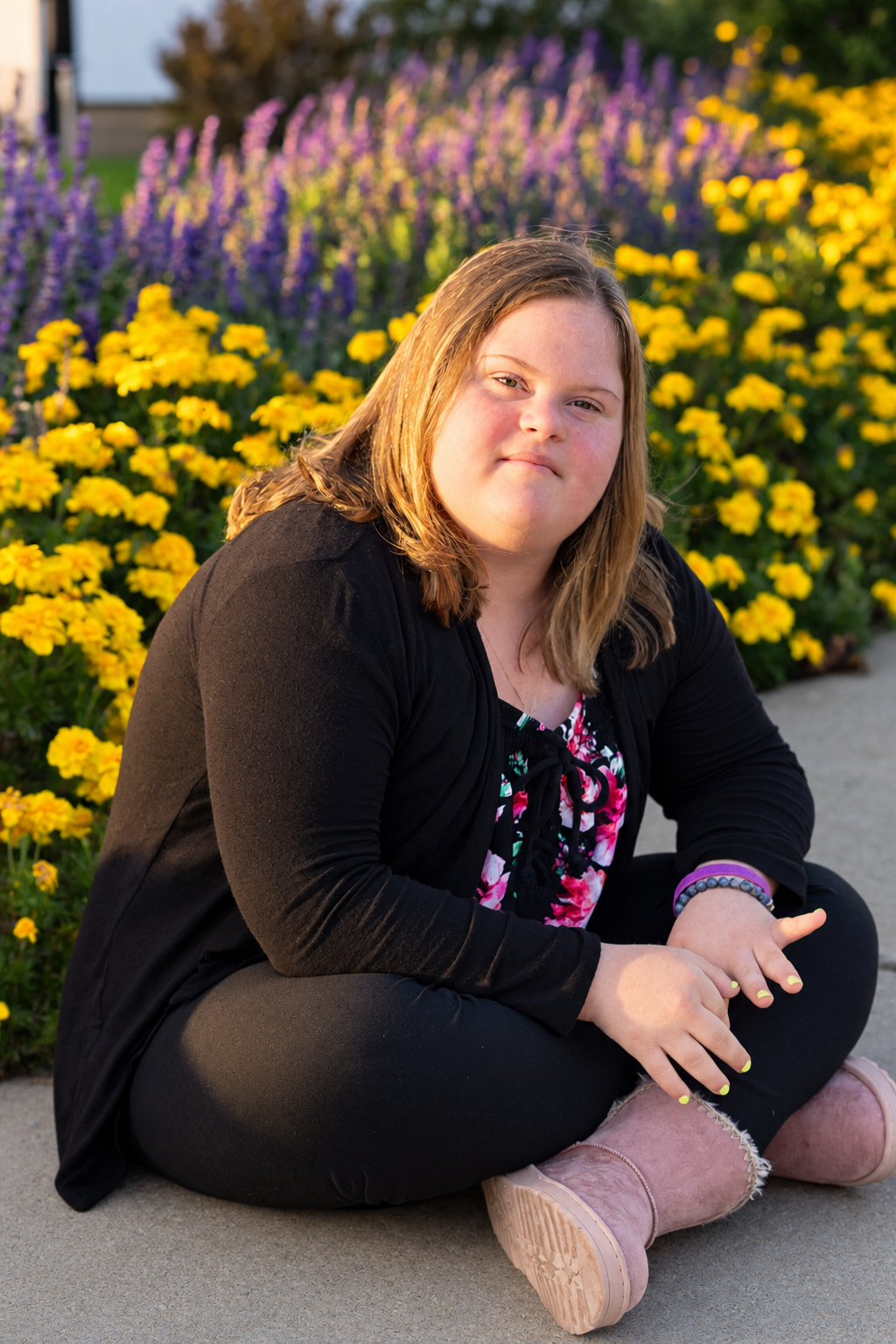 Young woman with Down syndrome sits cross-legged outdoors, smiling, with yellow and purple flowers blooming behind her.