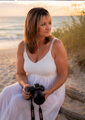 Woman in white dress holding a Canon camera sits on beach at golden hour sunset.