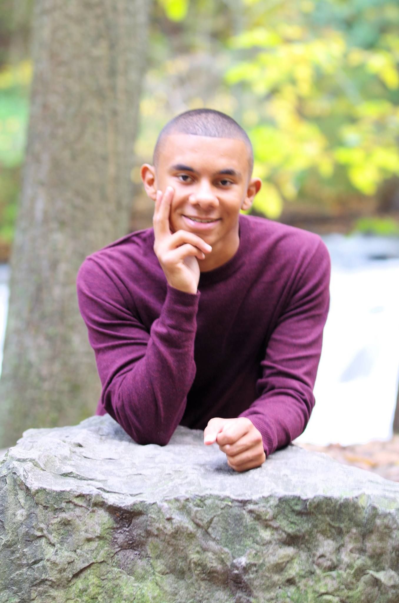 Student in burgundy sweater rests on stone ledge in outdoor nature setting.