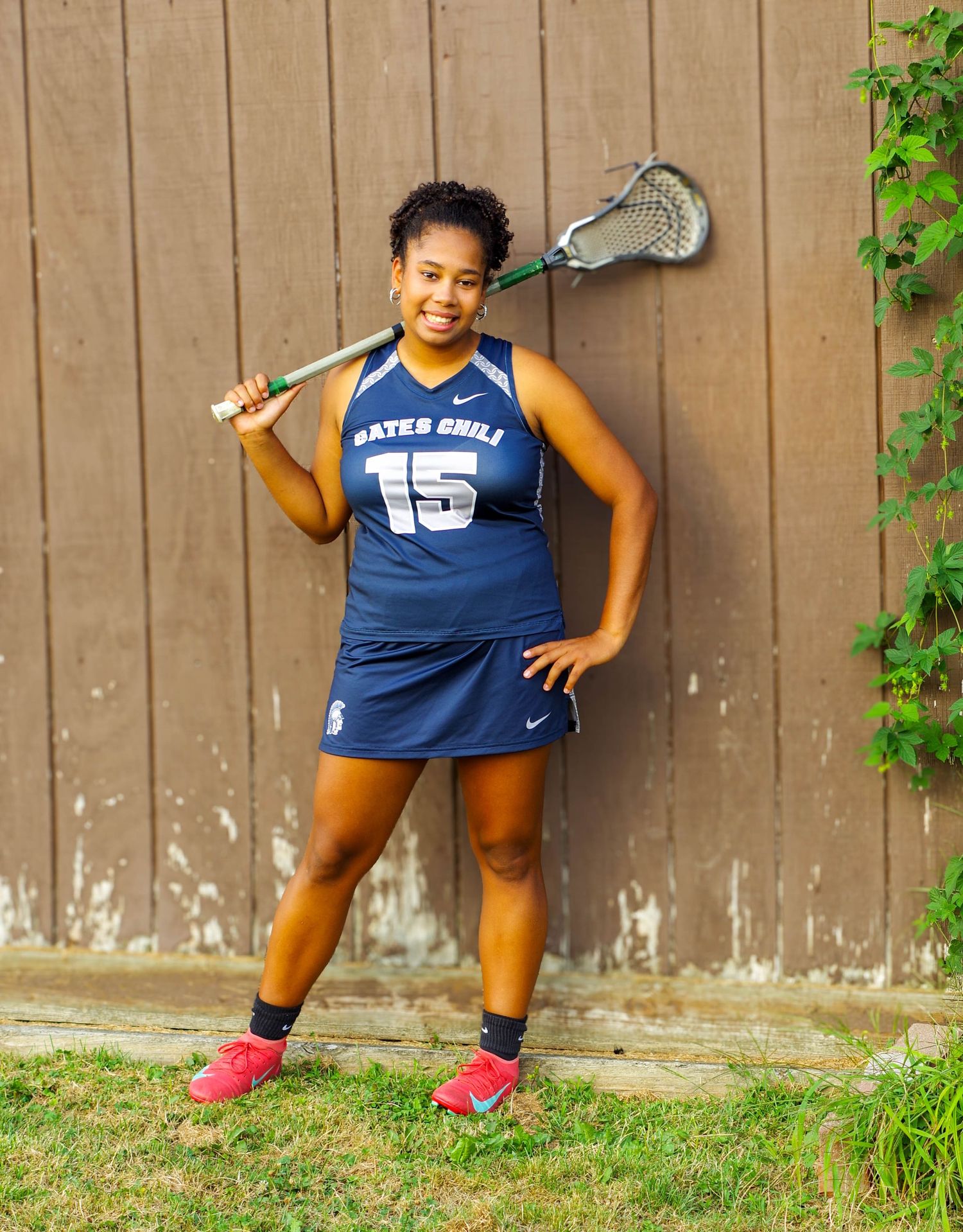 A lacrosse player in a blue team jersey poses against a brown fence with green vines.