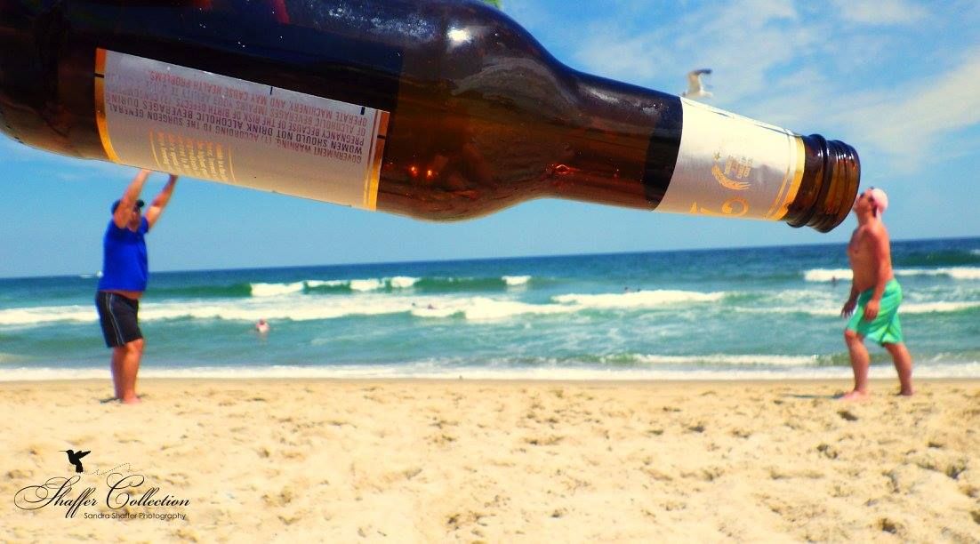 Beer bottle photographed on sandy beach with ocean waves in background.