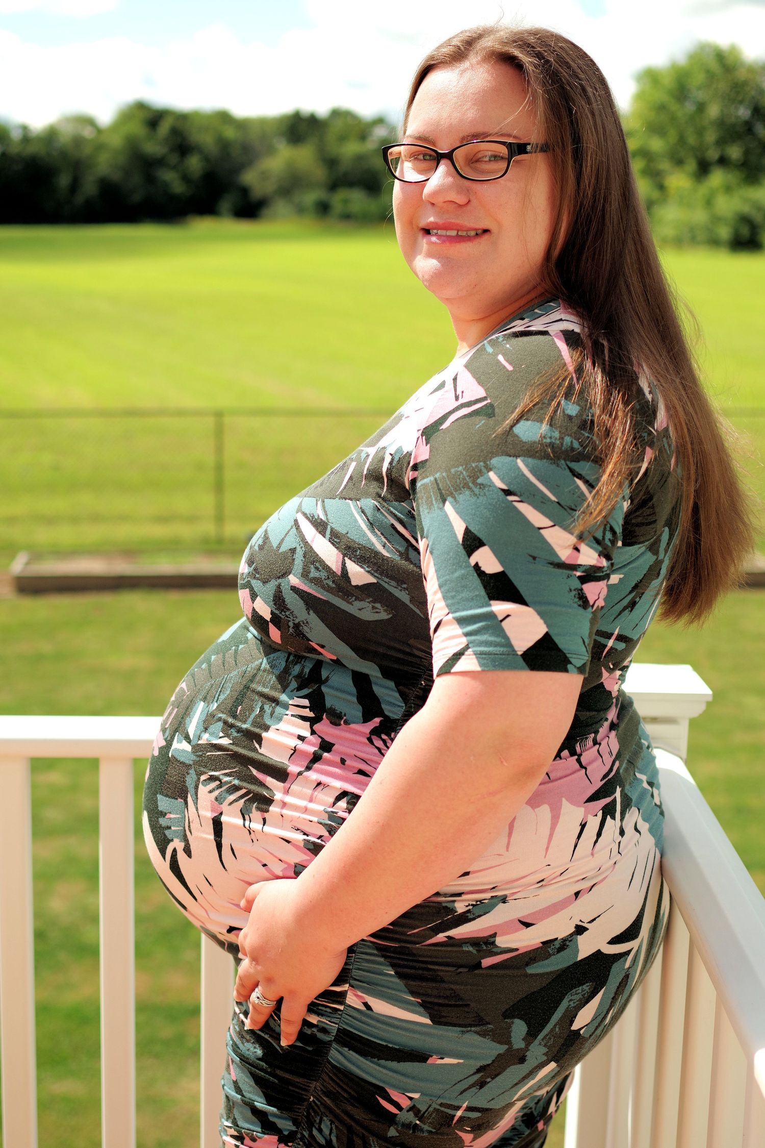 Maternity portrait on deck railing with sunny green background.