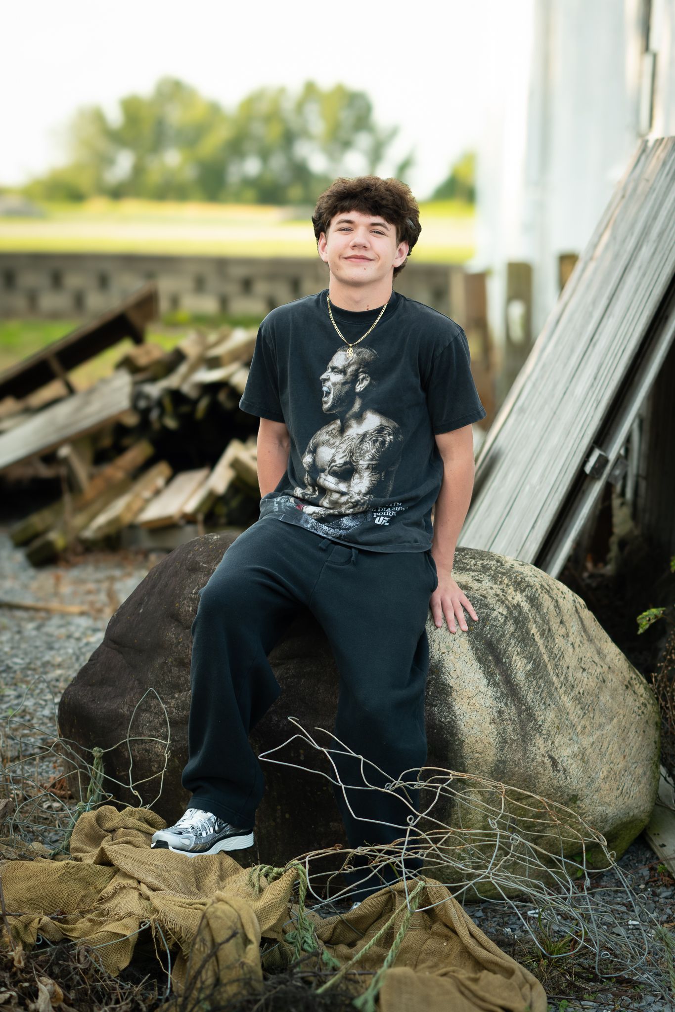 Teen boy in black graphic tee sits on large rock outdoors, smiling confidently near rustic wooden boards.