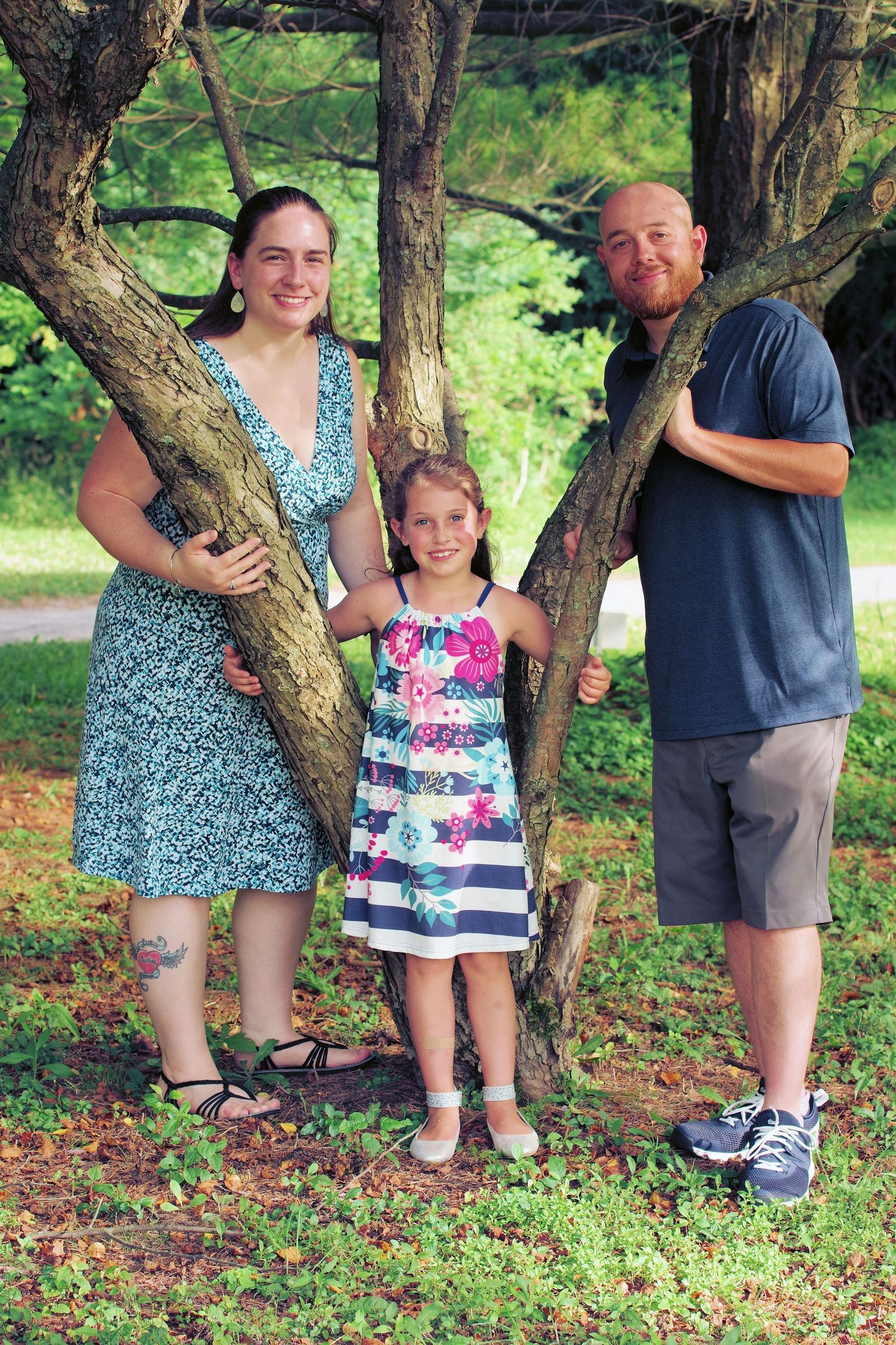 Family of three poses playfully around a tree trunk in a park setting with lush greenery.