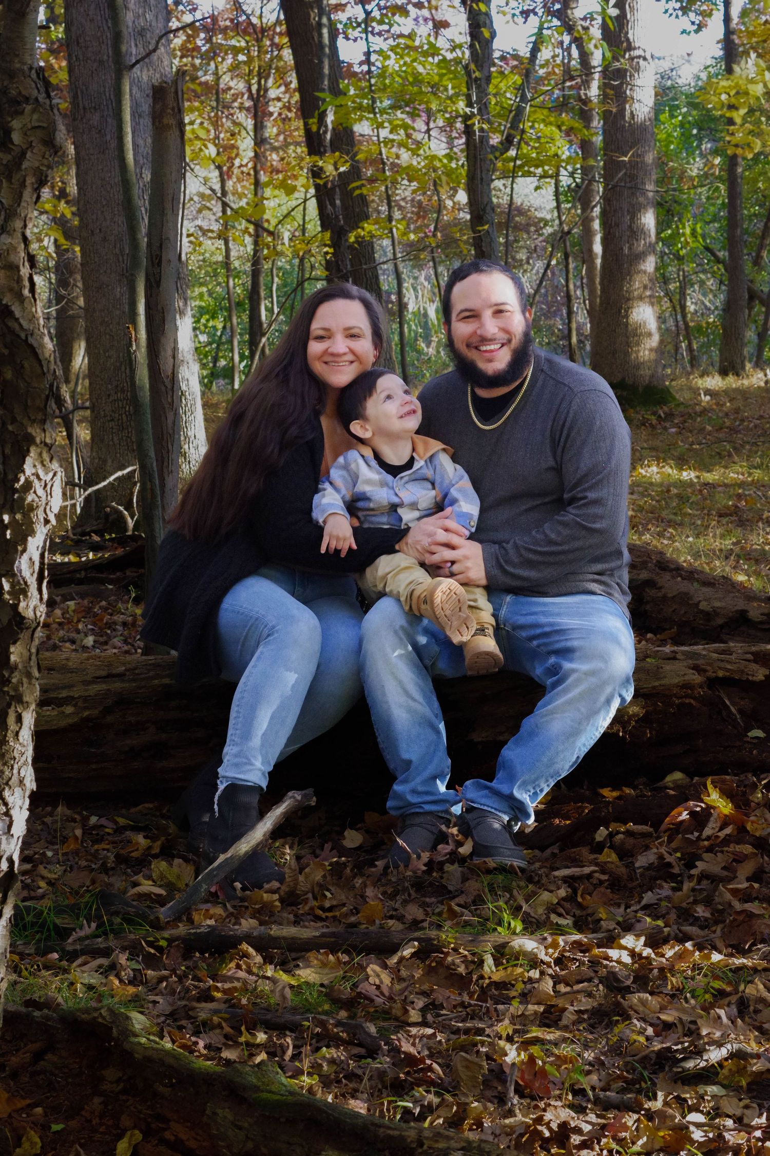 Family of three smiling on a fallen log in an autumn forest, parents holding a young toddler with fall foliage around them.
