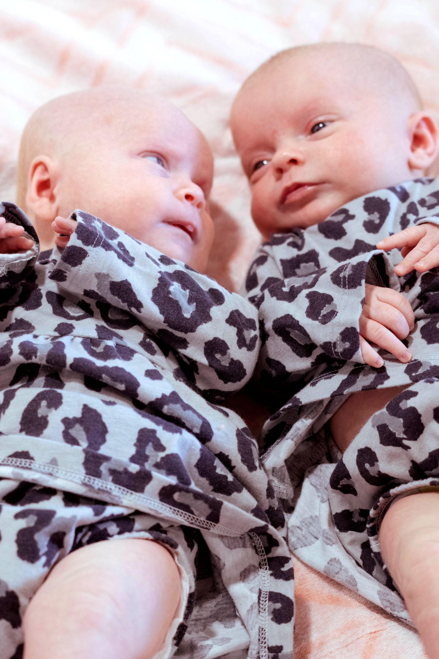 Two newborn twins in matching gray leopard print onesies lying side by side on a pink blanket.