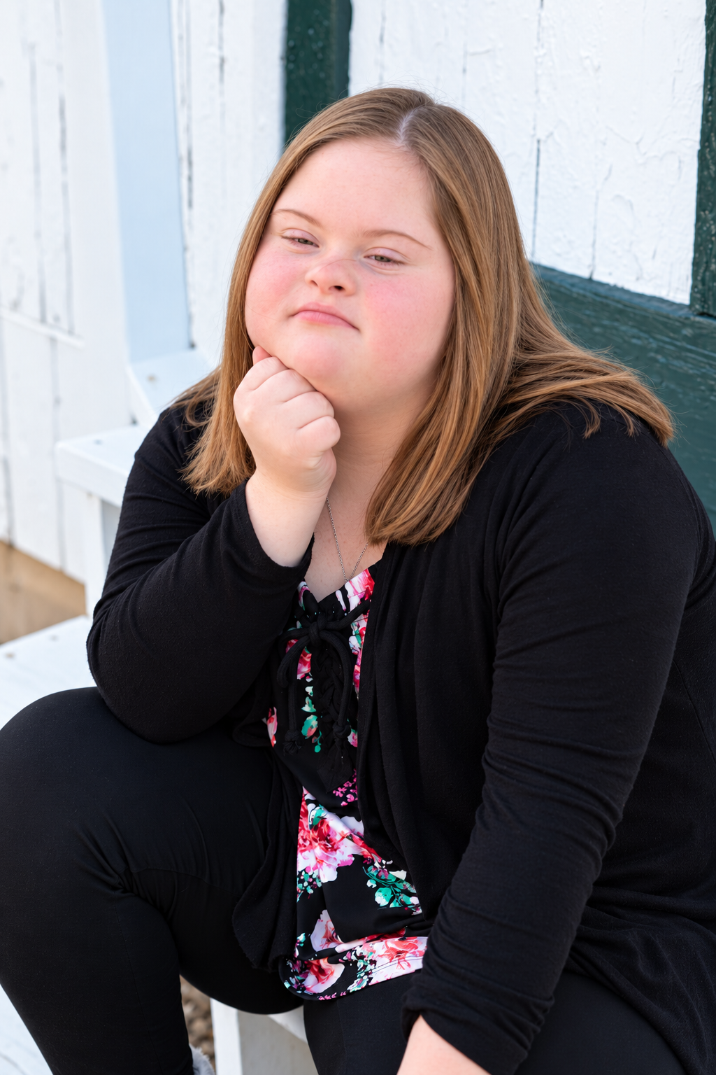Young girl with Down syndrome sitting outdoors, resting chin on fist in thoughtful pose, wearing black cardigan.