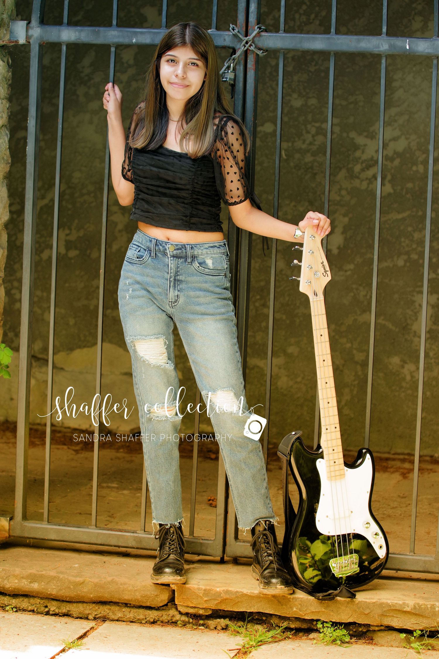 Musician poses with electric guitar against iron gate wearing black lace top and ripped jeans.
