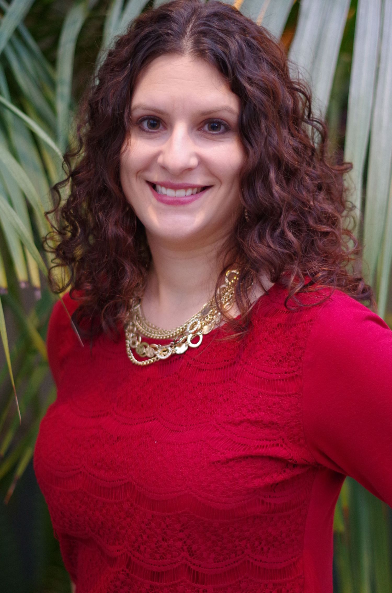 Professional portrait showing flowing dark curly hair, red sweater and gold statement necklace against a tropical background.
