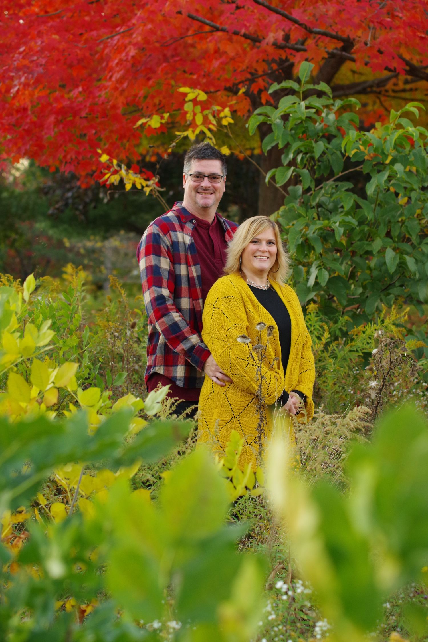 Couple standing together against vibrant red autumn foliage and green plants.