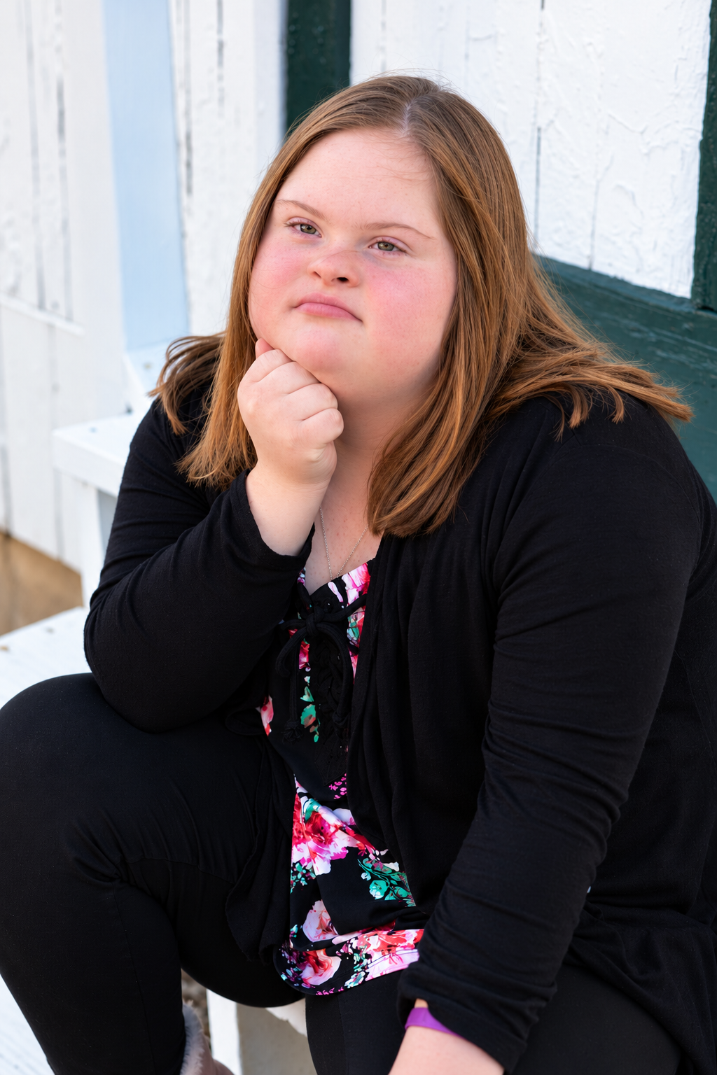 Young woman with Down syndrome in black cardigan and floral top sits thoughtfully with hand on chin outdoors.