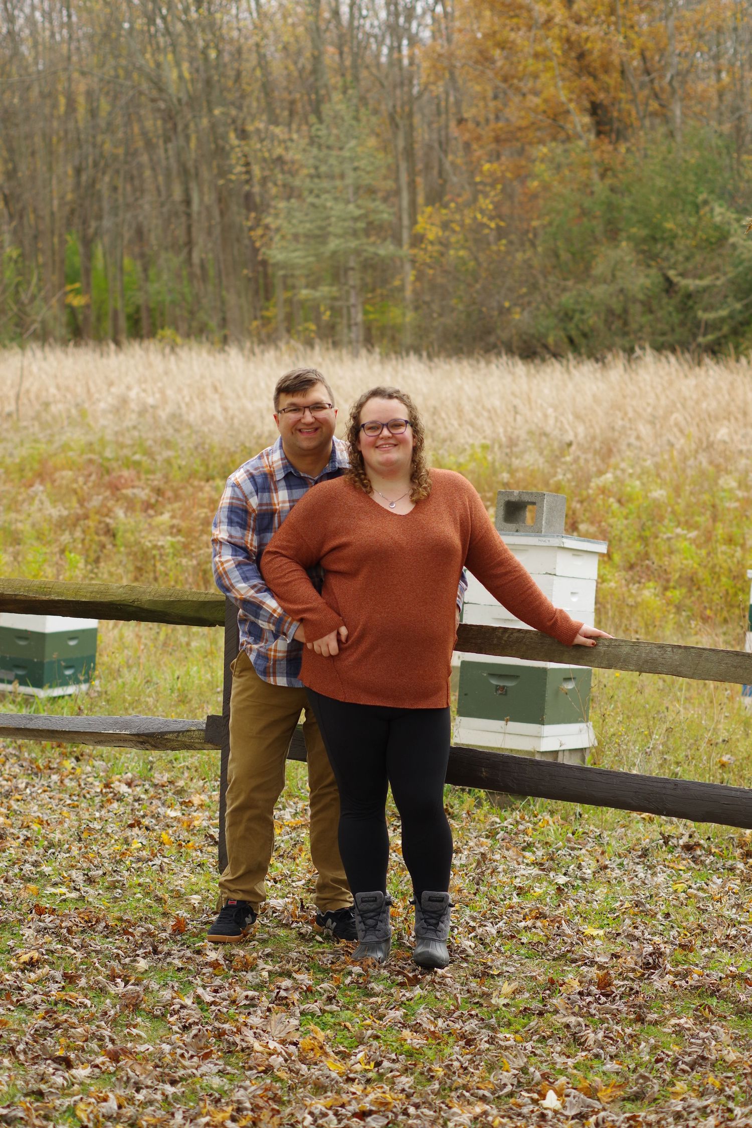 Couple standing by wooden fence in autumn field with trees and tall grass in background.