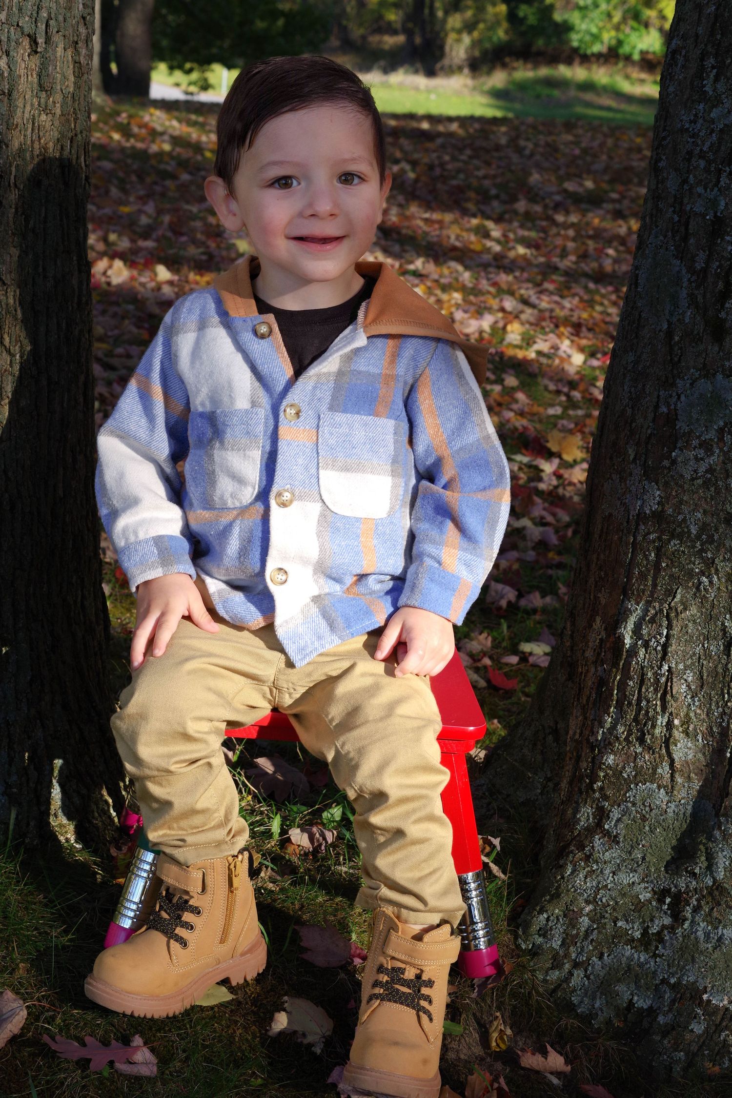 Smiling child wearing plaid button-up shirt and khaki pants seated between trees in autumn leaves.