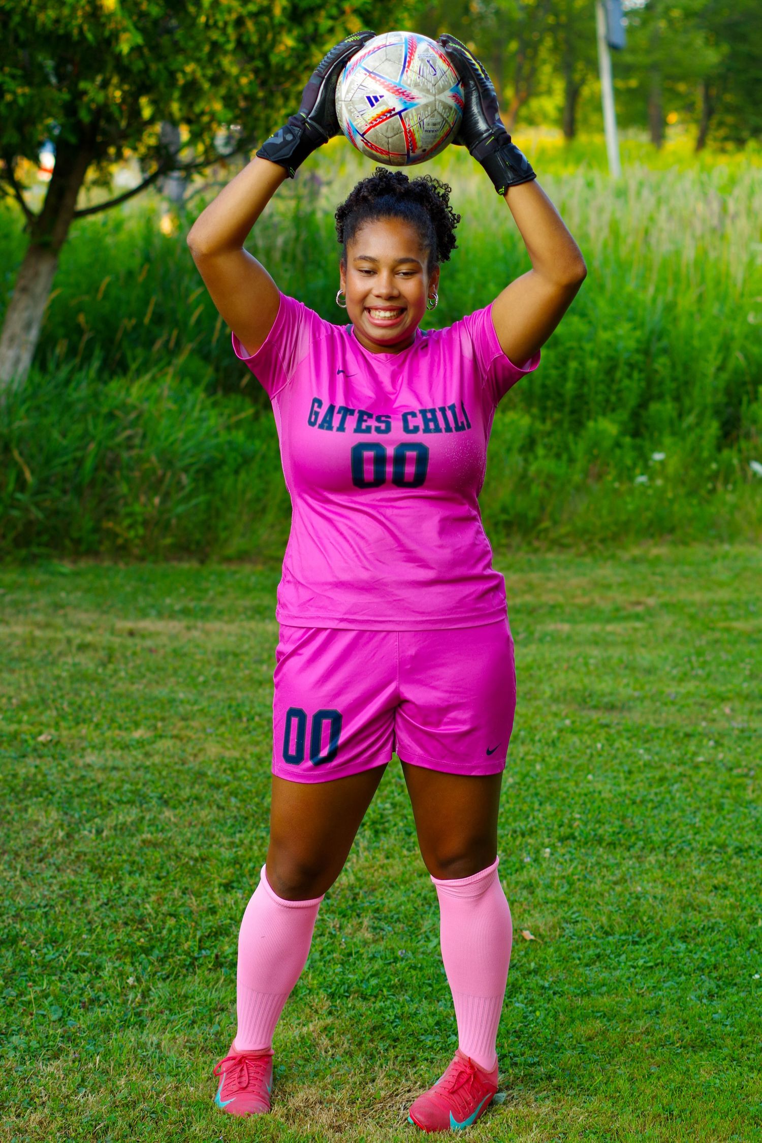 Soccer player in pink uniform holds ball high while smiling on green grass field during practice session.