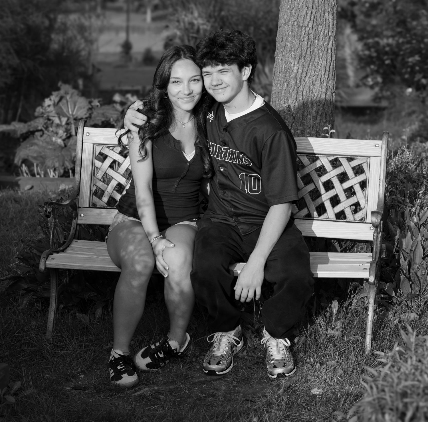Black and white photo of a smiling young couple sitting together on a wooden bench outdoors near a tree.