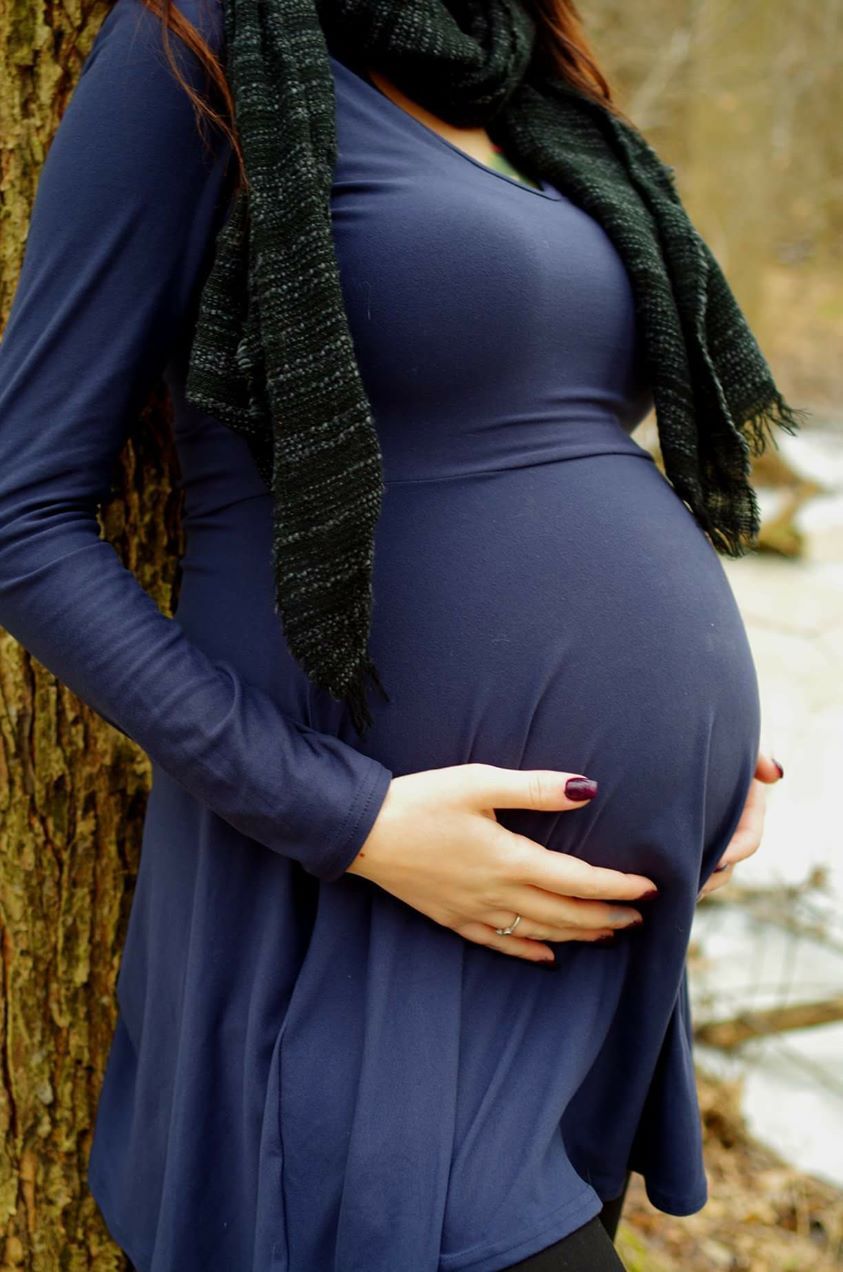 Pregnant woman in blue dress and scarf cradling baby bump outdoors near tree in winter.