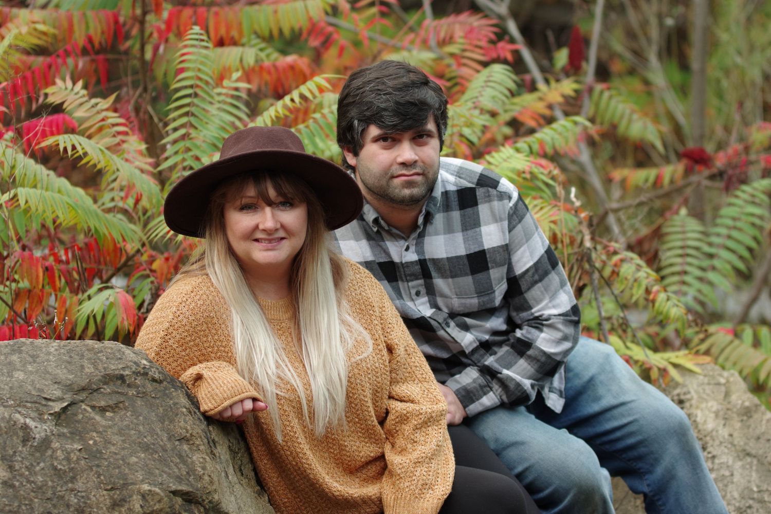 Couple posing on rocks surrounded by vibrant red and green autumn foliage, woman in orange sweater and hat.