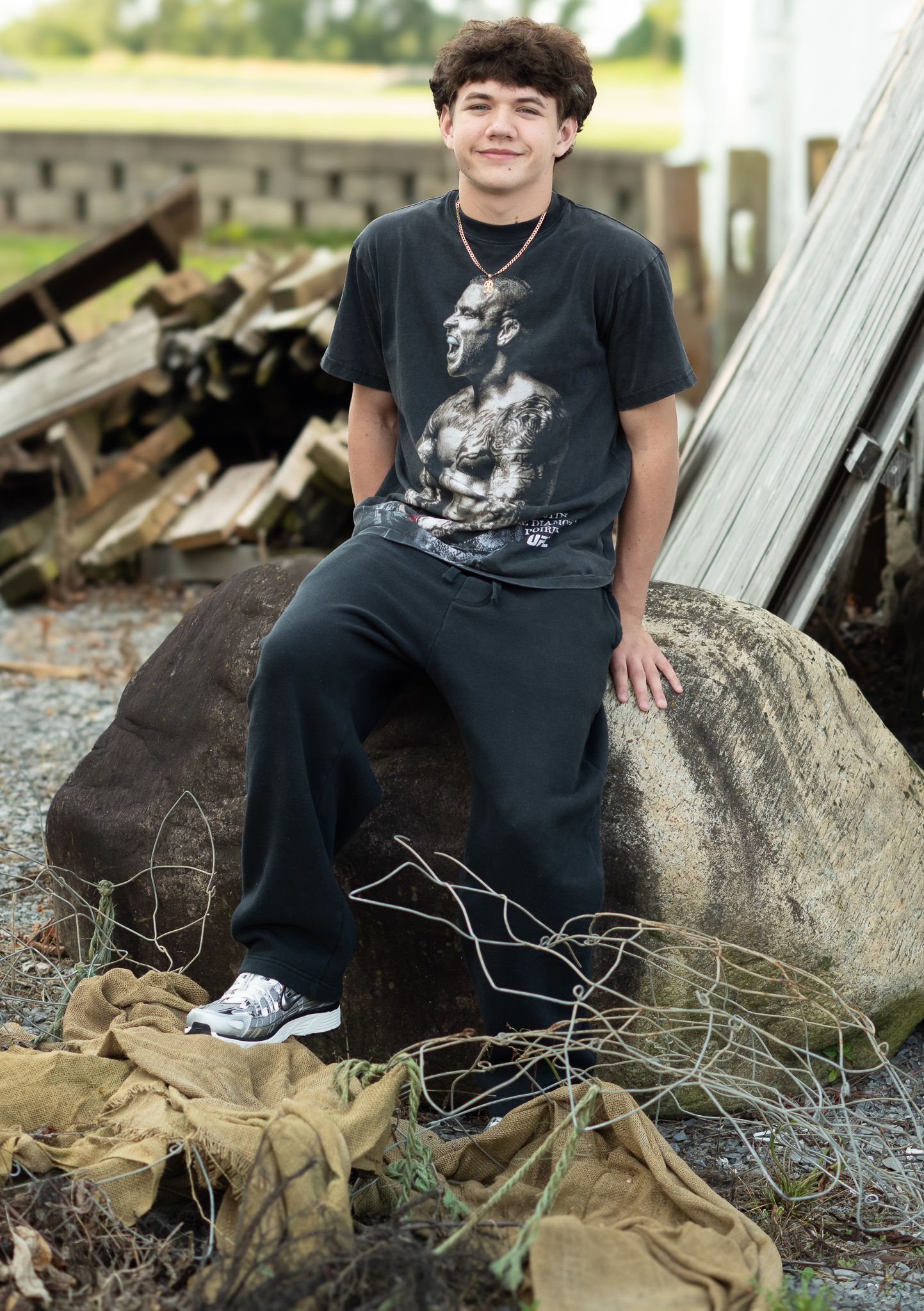 Teen boy in black graphic tee and joggers sits on a large rock outdoors near wooden pallets.