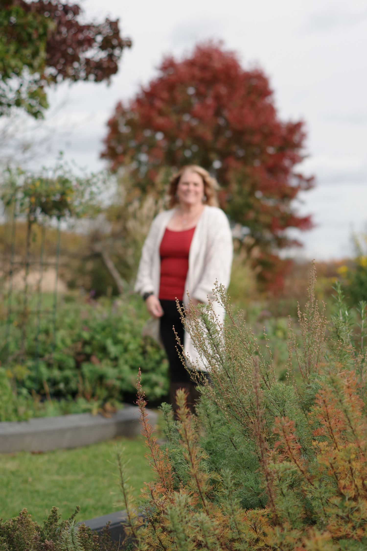 Person in red top and cream cardigan standing in garden with tall plants.