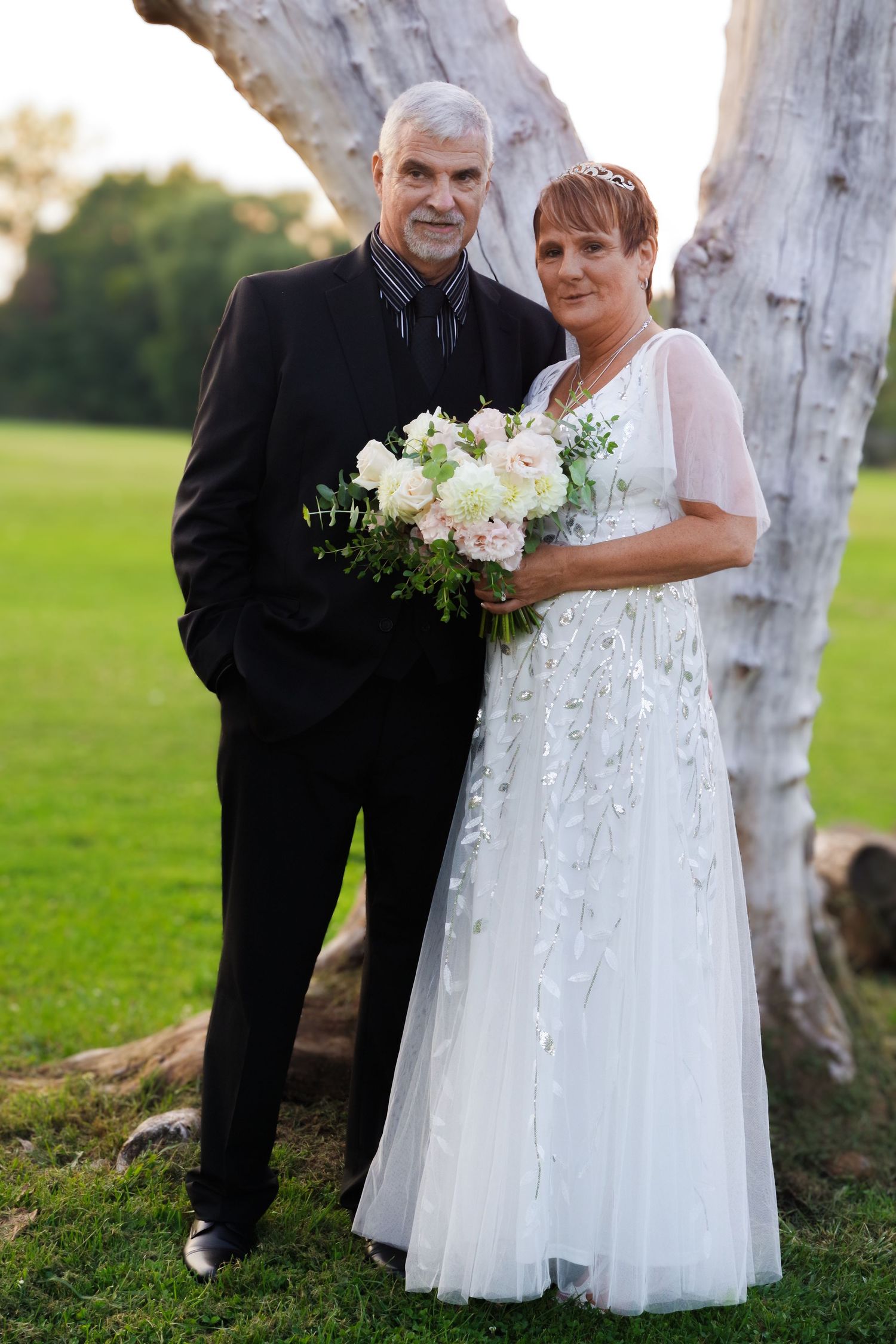A couple in formal wedding attire stands by a white tree trunk holding a bouquet.