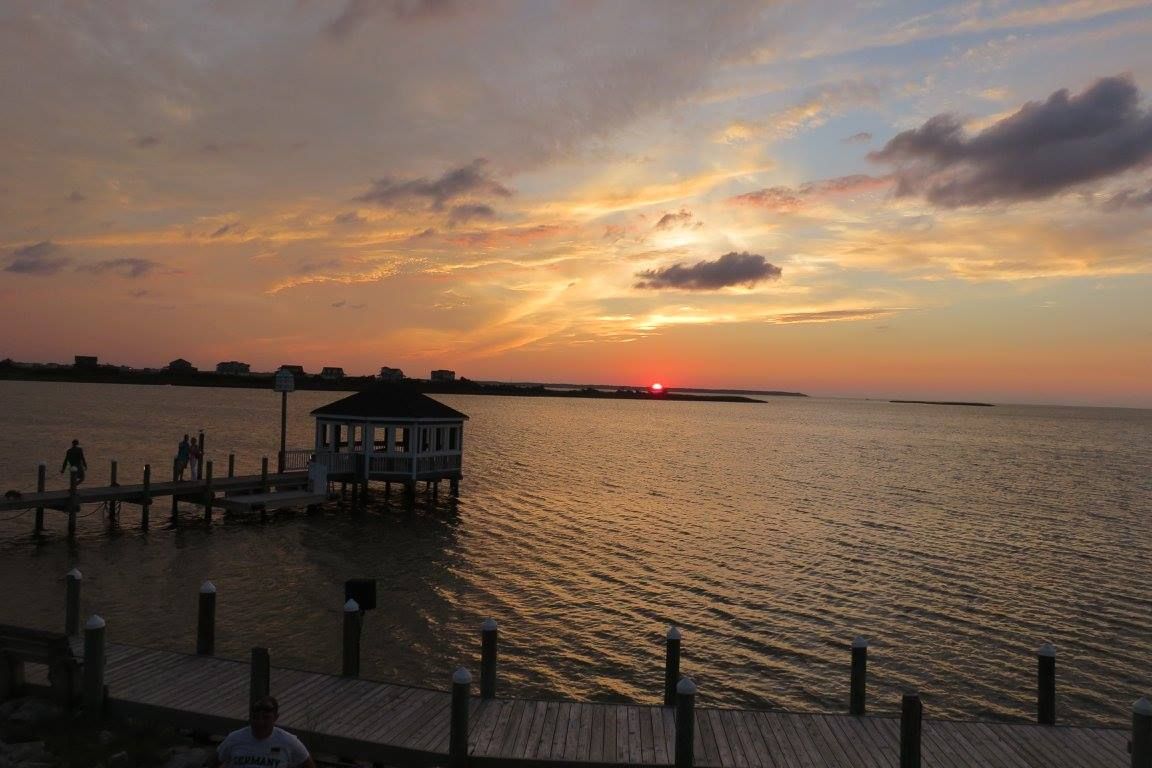 A wooden dock extends over calm waters during a dramatic orange sunset with a small fishing hut silhouetted against the sky.
